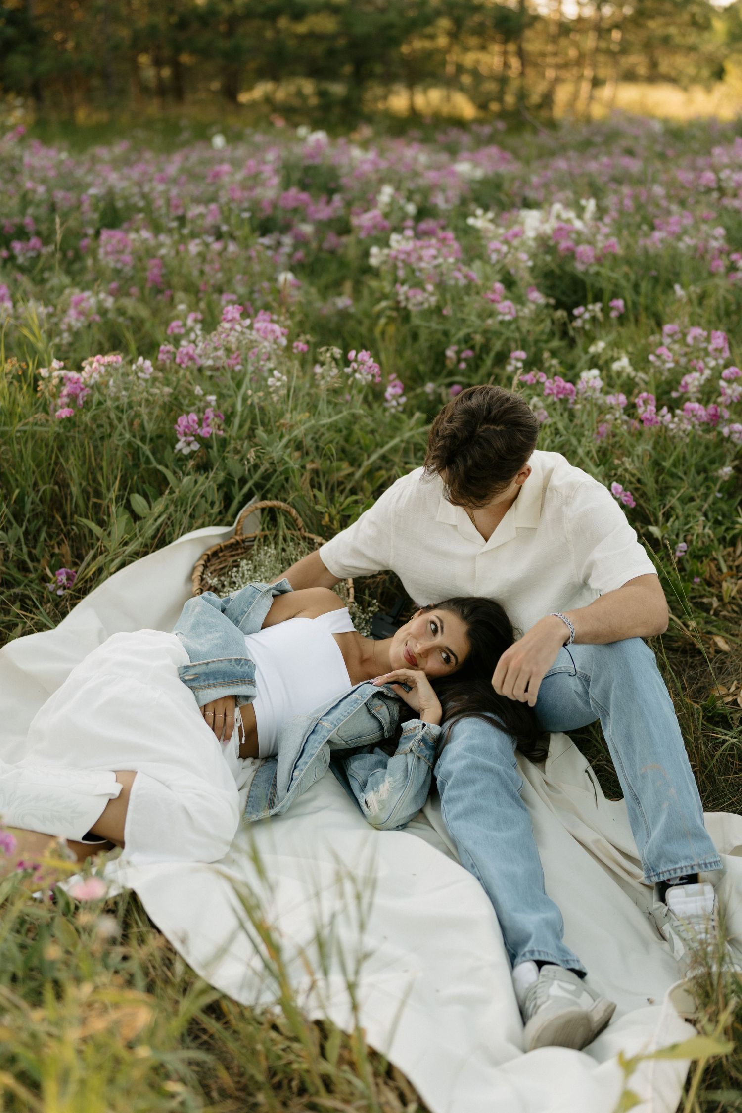 Couple lying on white blanket surrounded by pink wildflowers during outdoor photoshoot.