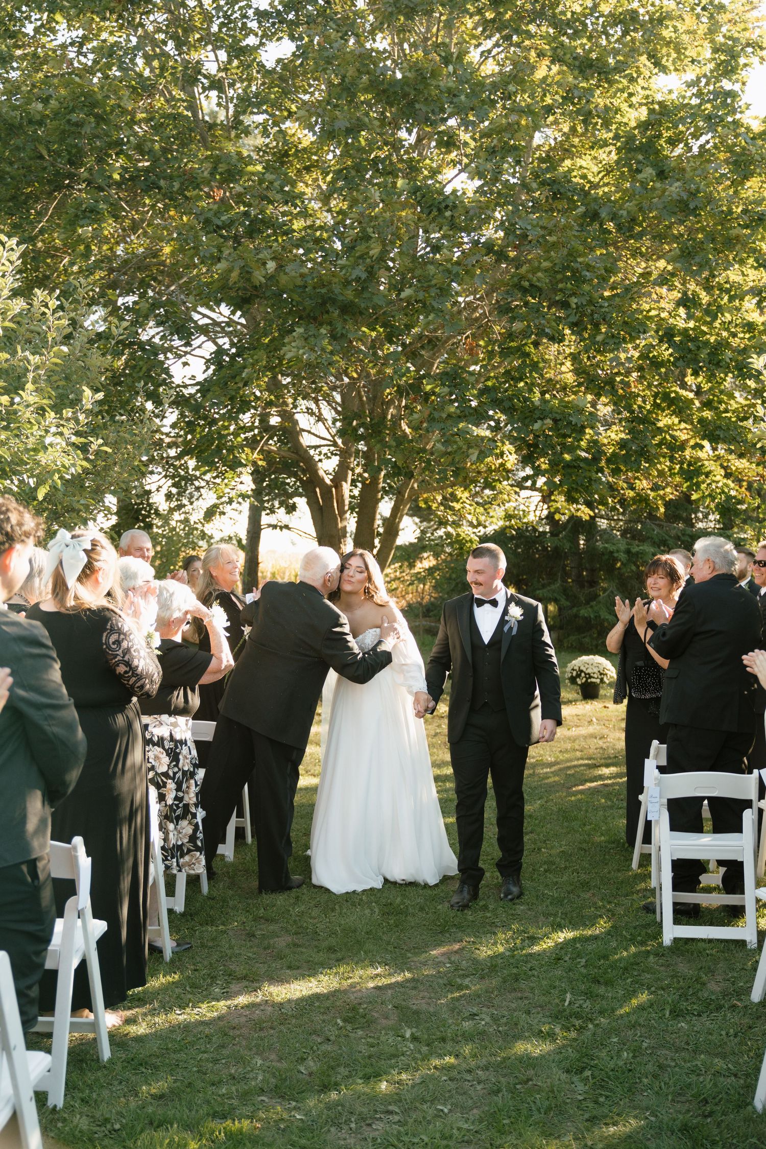 Outdoor wedding ceremony under large tree with guests seated on white chairs.