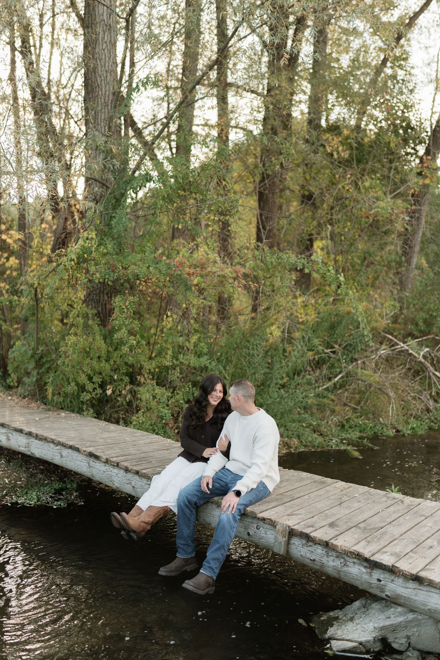 A couple sits together on a wooden dock over a peaceful pond surrounded by trees.