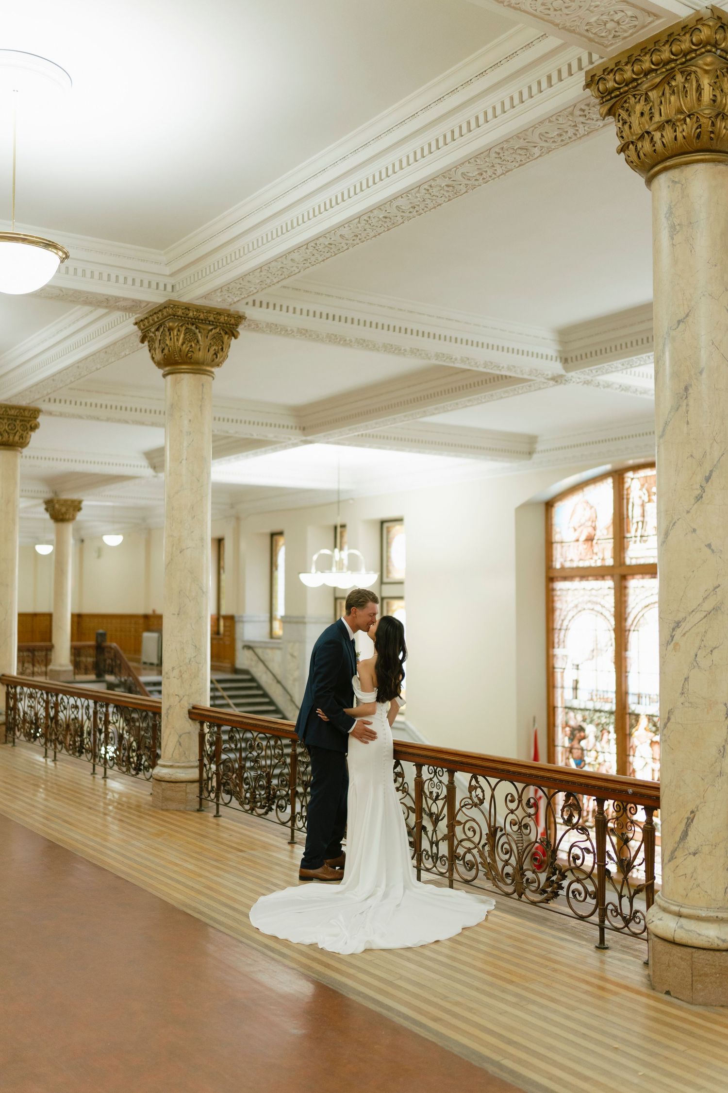 Bride and groom share a romantic kiss on an ornate staircase landing with marble columns in an elegant historic building.