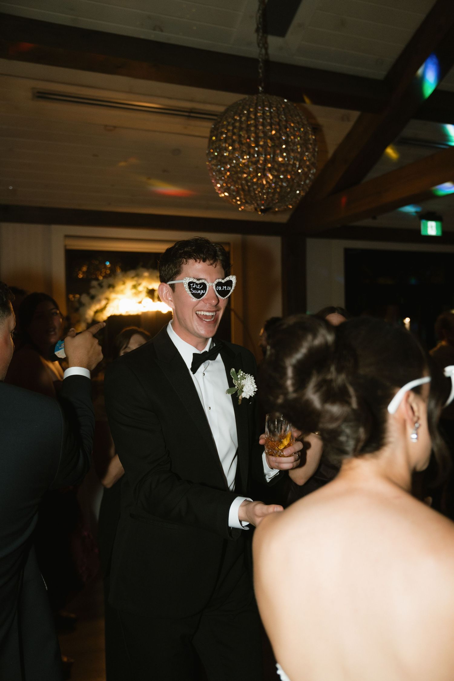 Groom in tuxedo wearing heart-shaped sunglasses smiling at wedding reception with chandelier backdrop.