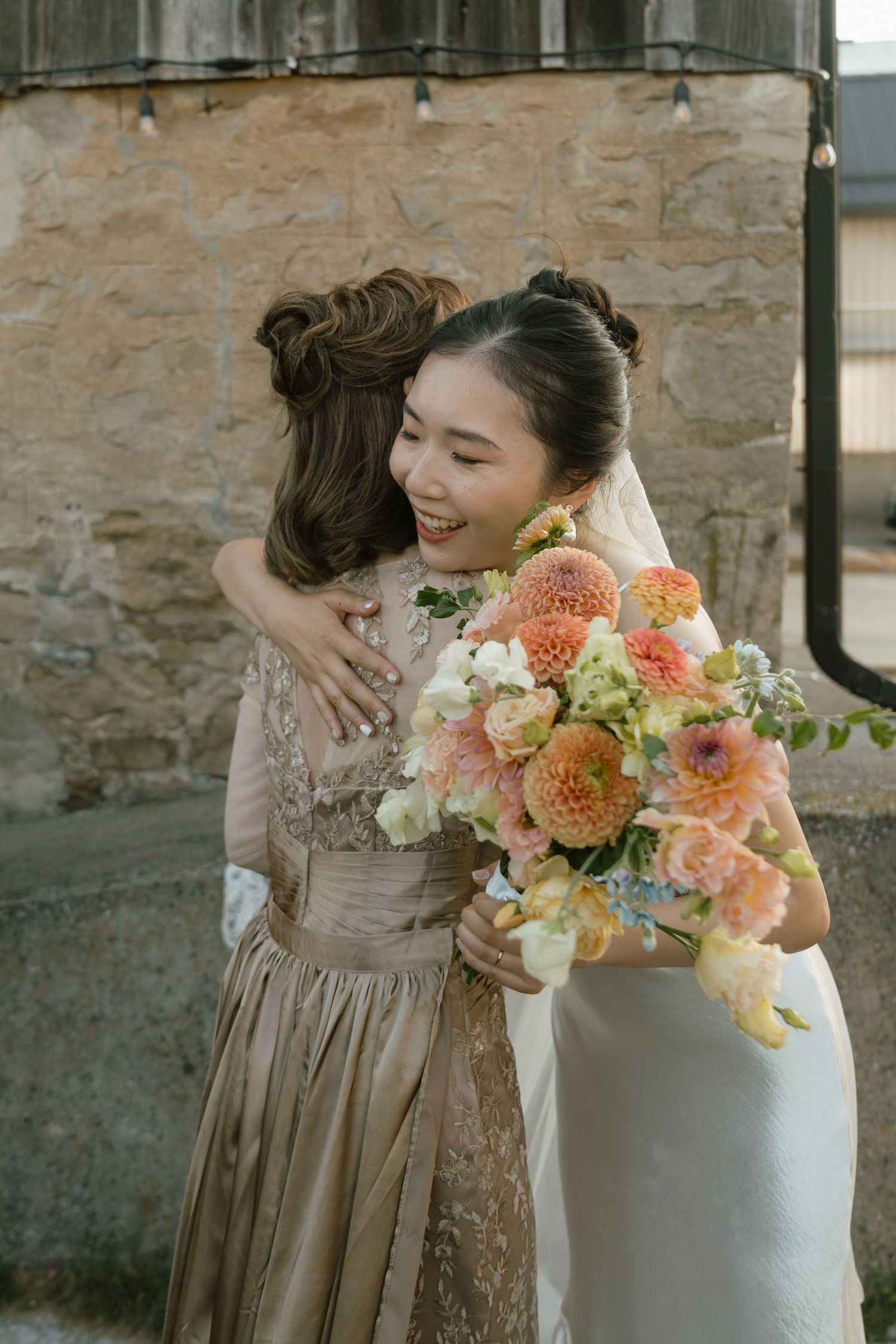 Wedding party embraces with peach and coral floral bouquets against stone wall.