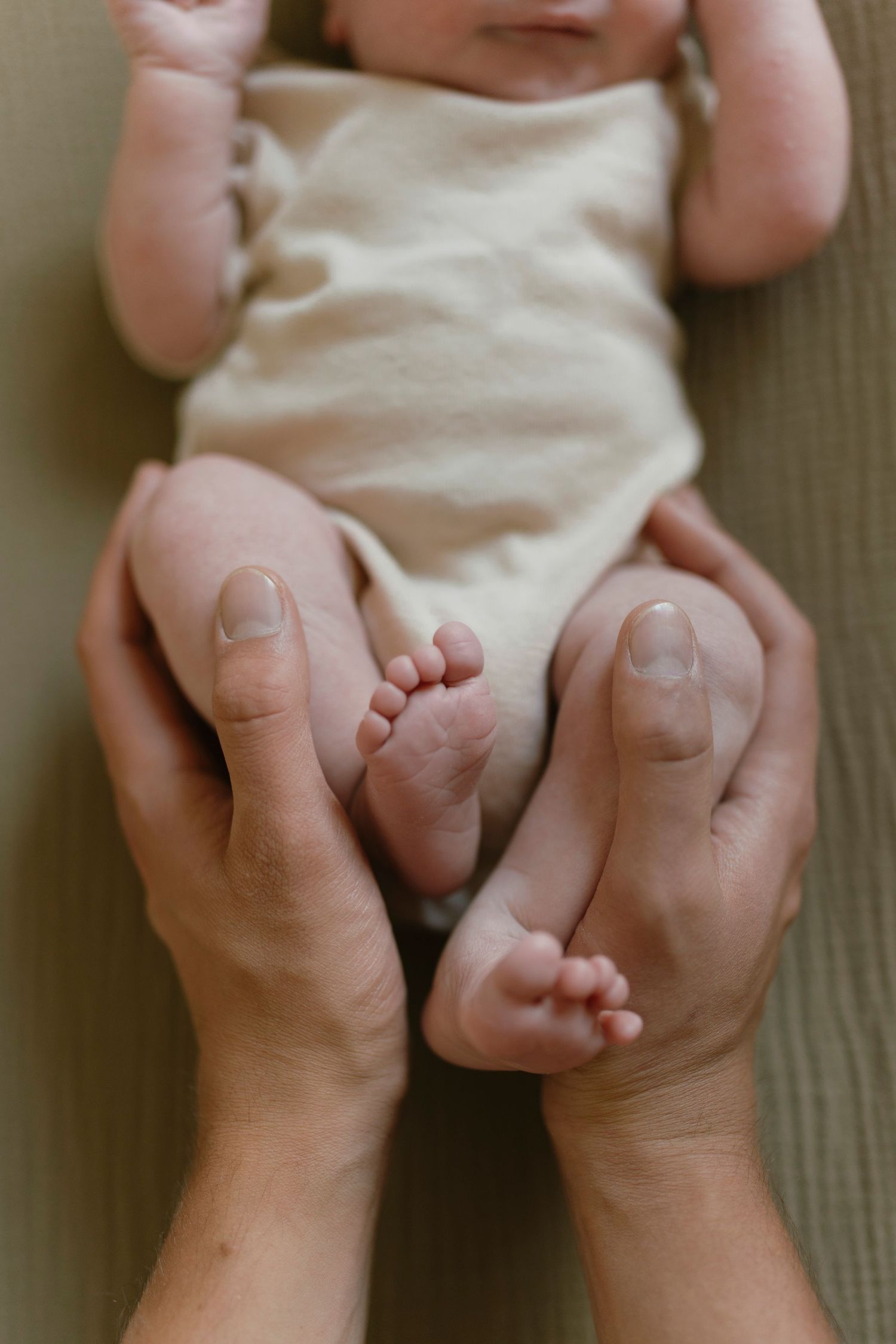 Adult hands gently cradling tiny newborn baby feet, with infant in cream onesie lying on soft surface.