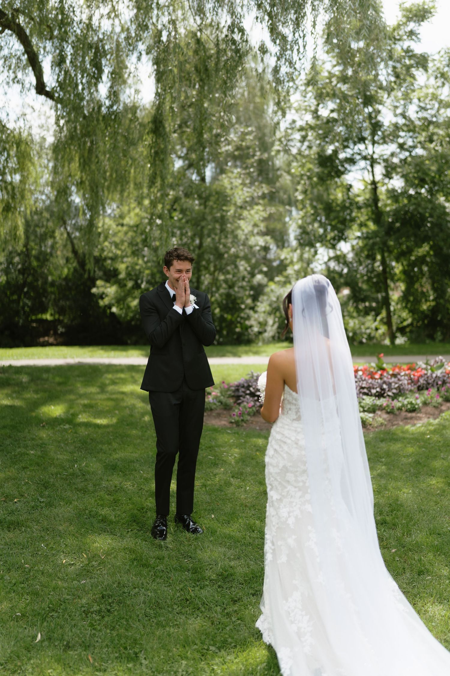 Groom in black tuxedo reacts emotionally during first look with bride in lace gown and veil in garden.
