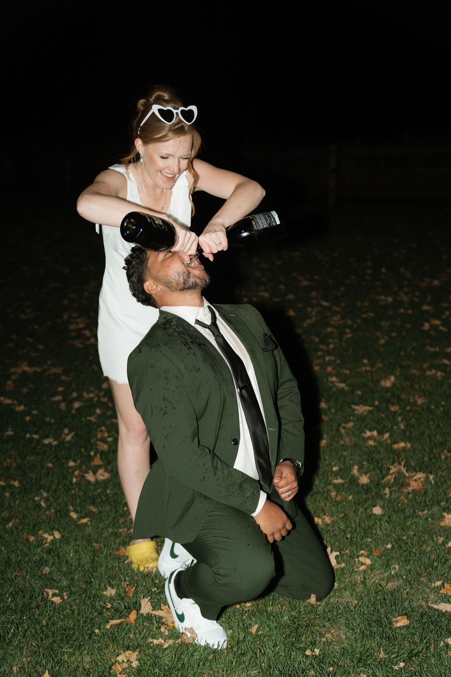 Two people playfully share drinks while sitting on grass at nighttime.
