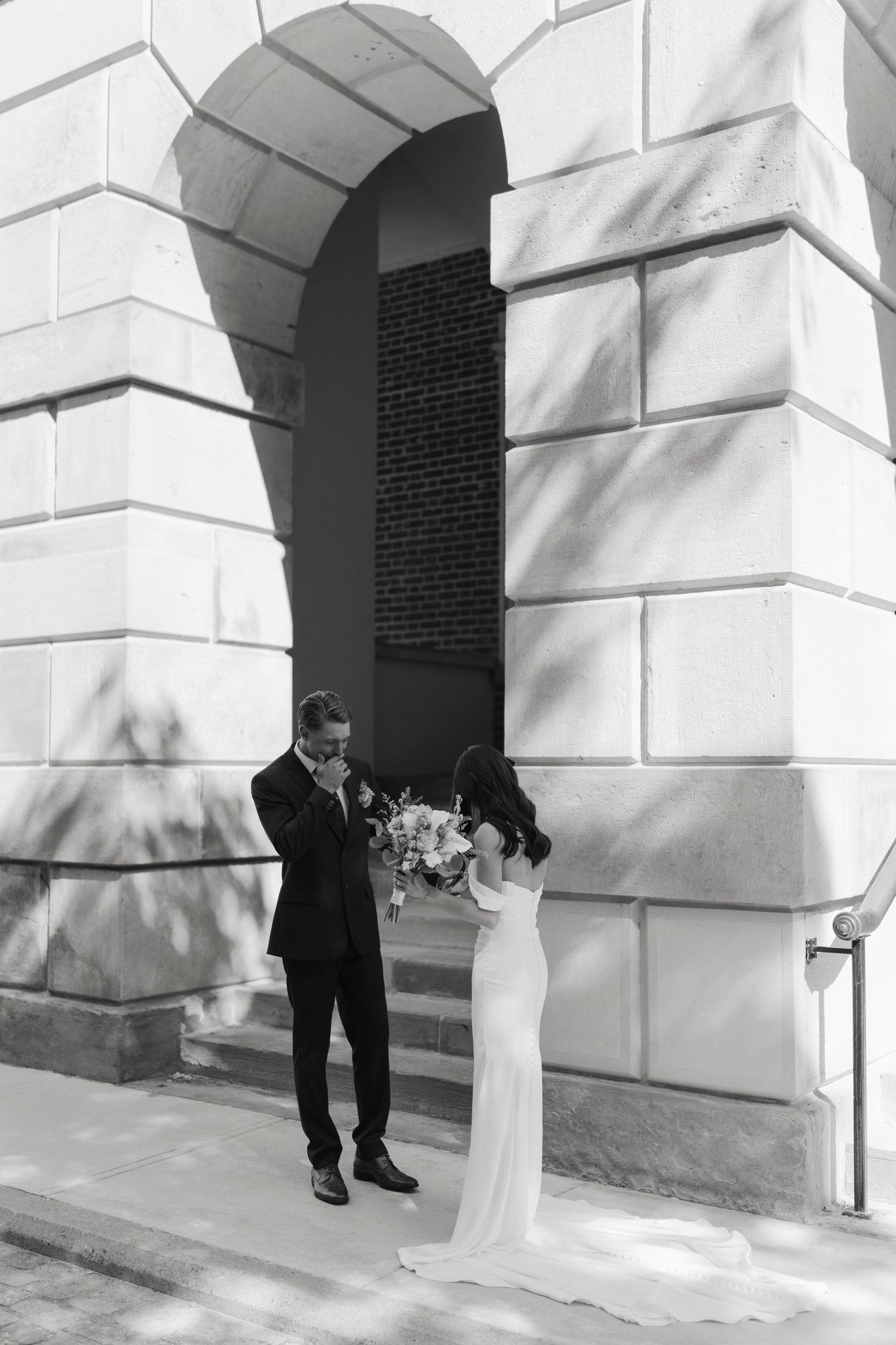 Black and white photo of a bride and groom sharing a tender moment outside a stone archway building.
