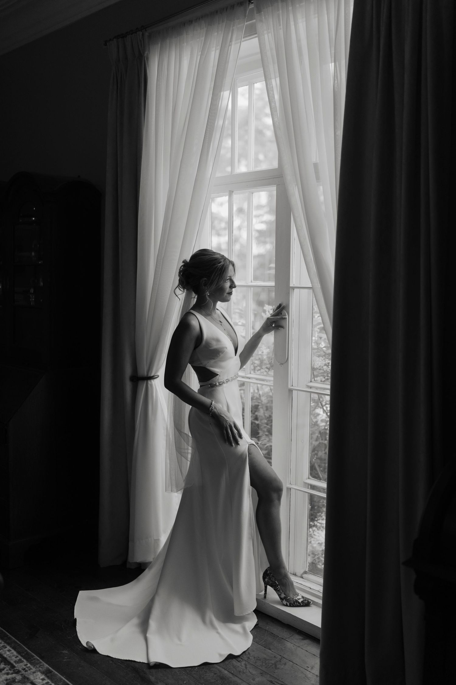 Black and white photo of a bride in a sleek wedding gown holding a glass, gazing out a large window with sheer curtains.