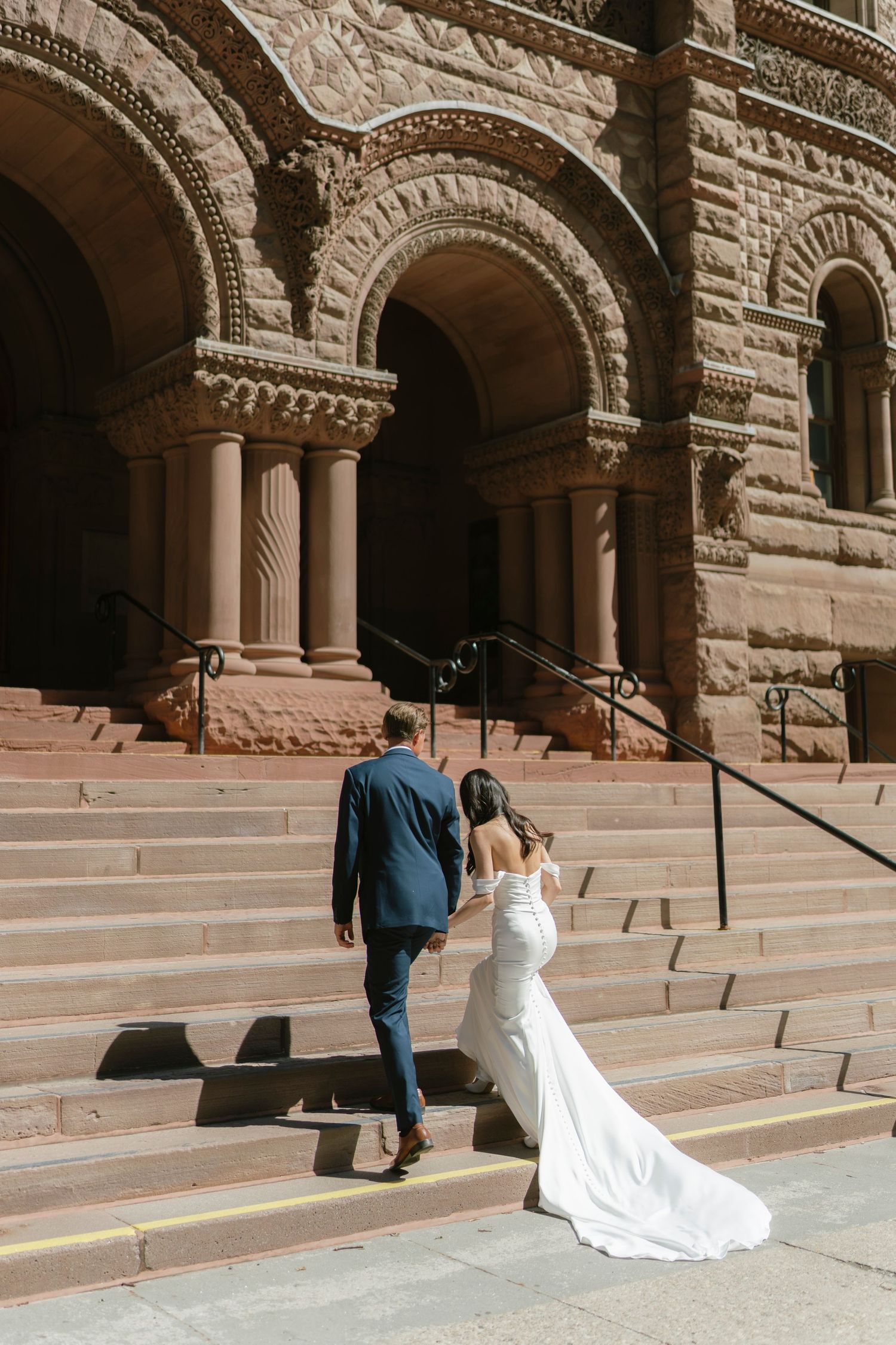 Bride and groom walking up stone steps of ornate historic building, bride in elegant white gown with train.