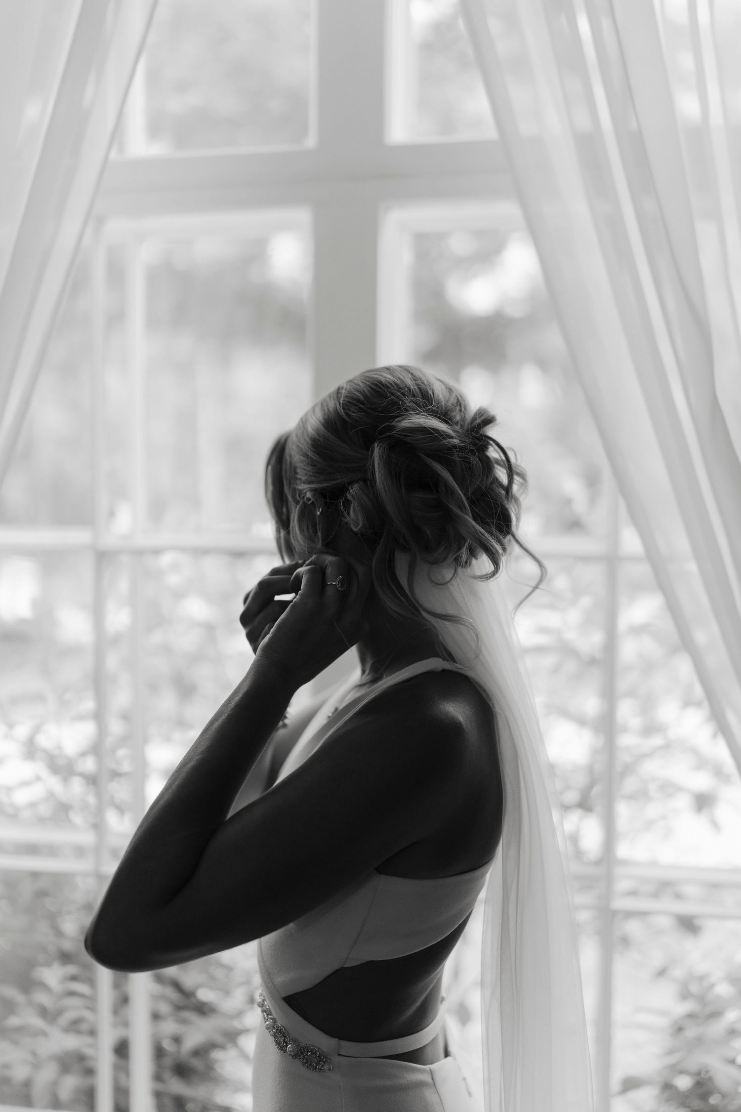 Black and white photo of a bride in lingerie and veil standing by a window, adjusting her earring.