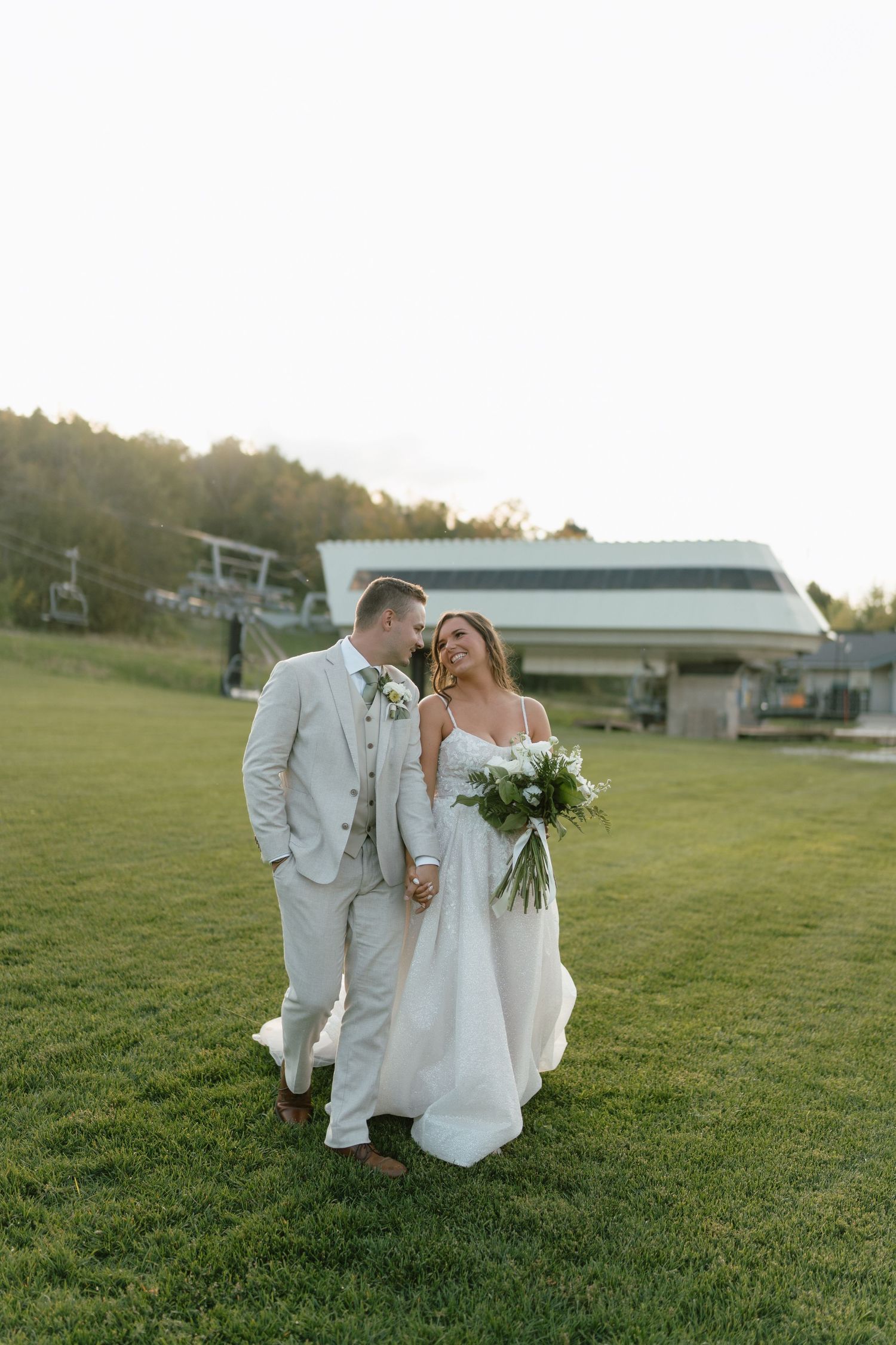 A couple in white attire shares an intimate moment on a grassy hillside.