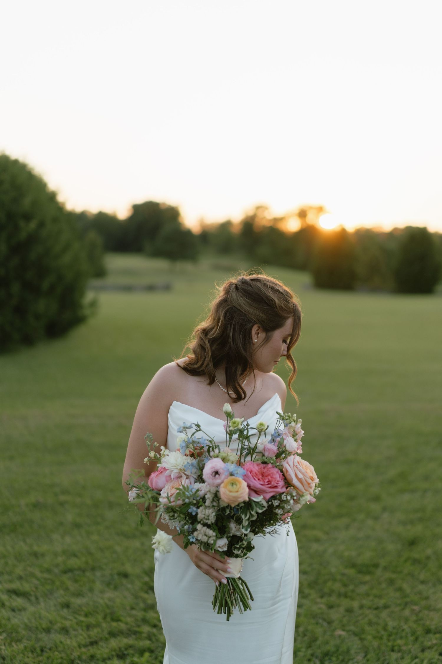 Bride in white strapless gown holds pink floral bouquet at golden hour in lush green field.