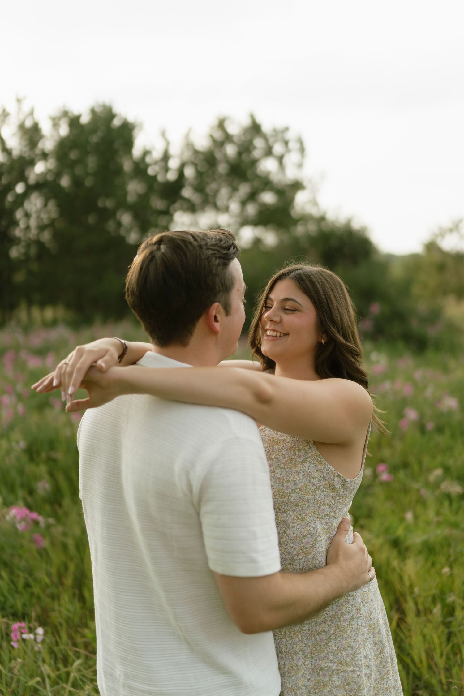 A couple embraces and smiles in a blooming wildflower field, the woman in a floral dress gazing lovingly at her partner.