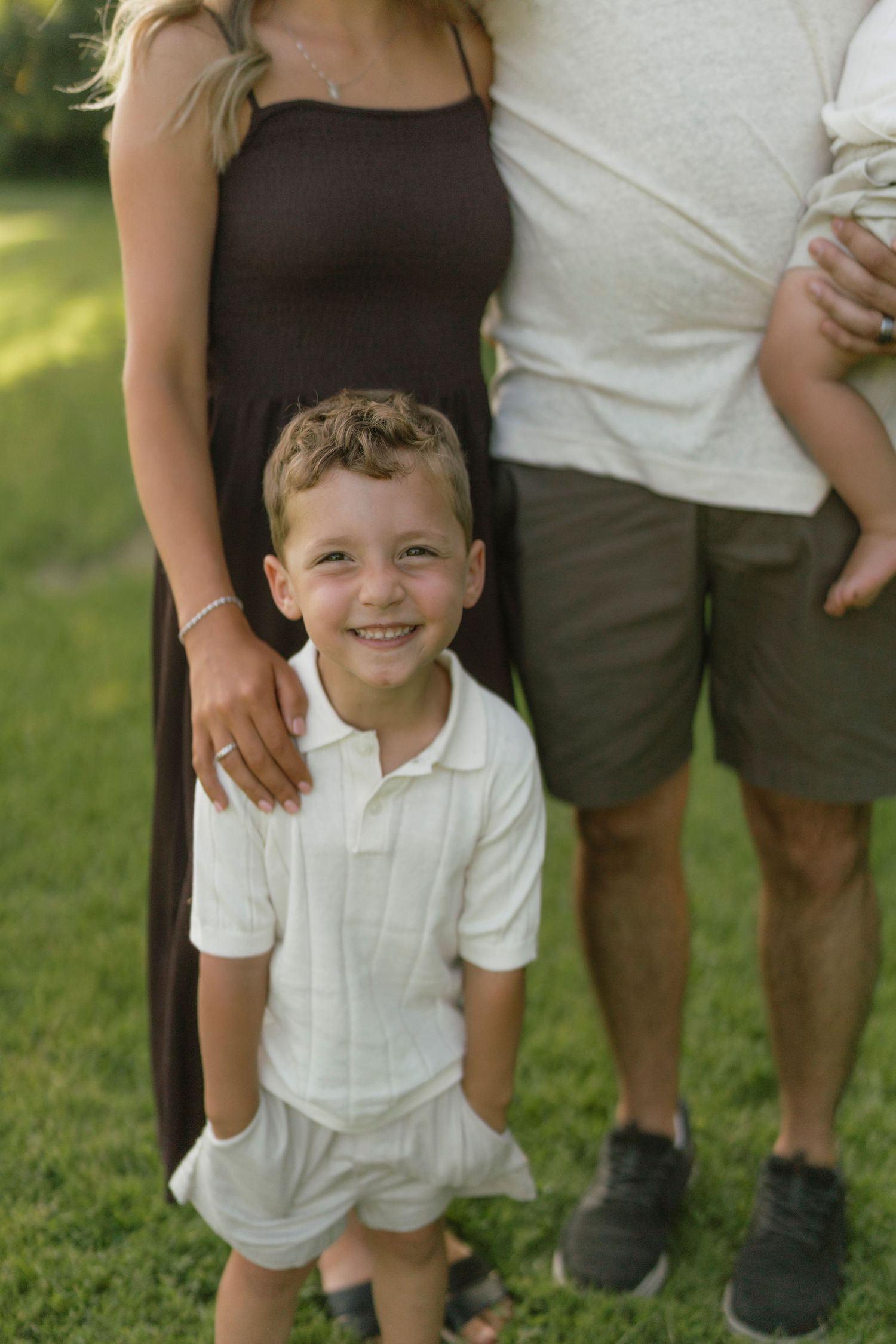 A smiling young boy in a white polo shirt stands in front of his parents on a green lawn.
