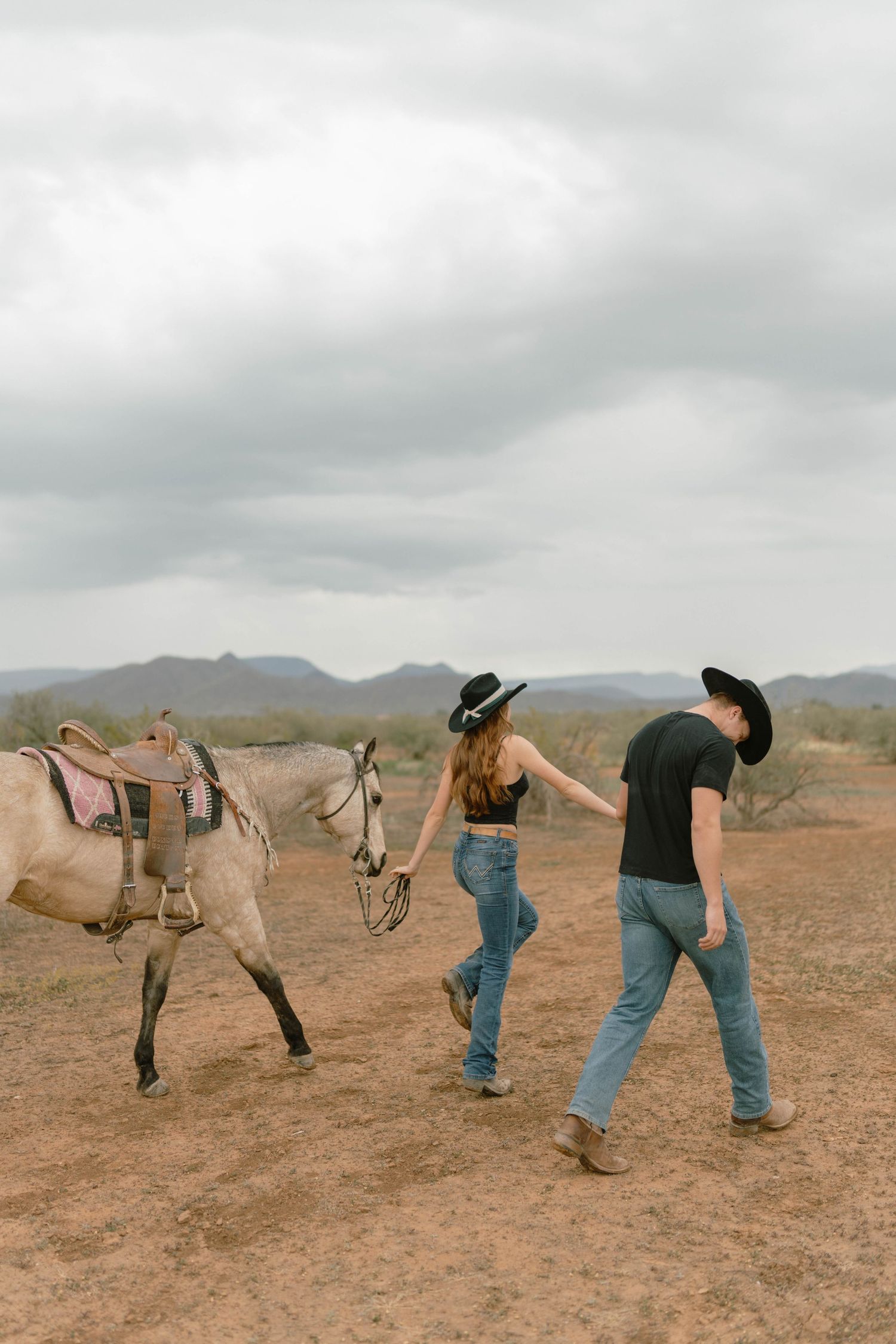 Couple in western wear walking with horse through desert terrain under cloudy skies.