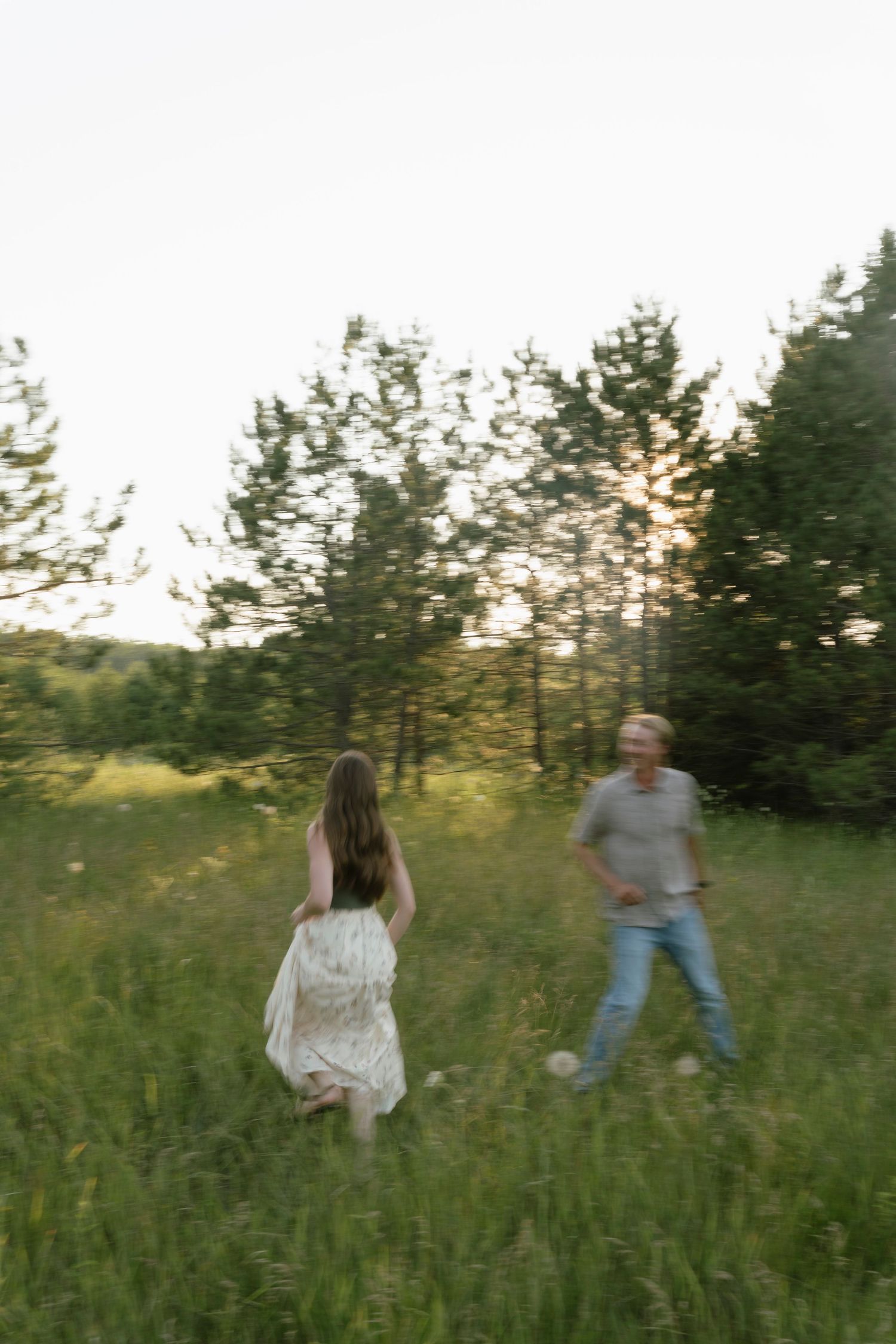 A couple walks through a sunlit green meadow surrounded by trees during golden hour.