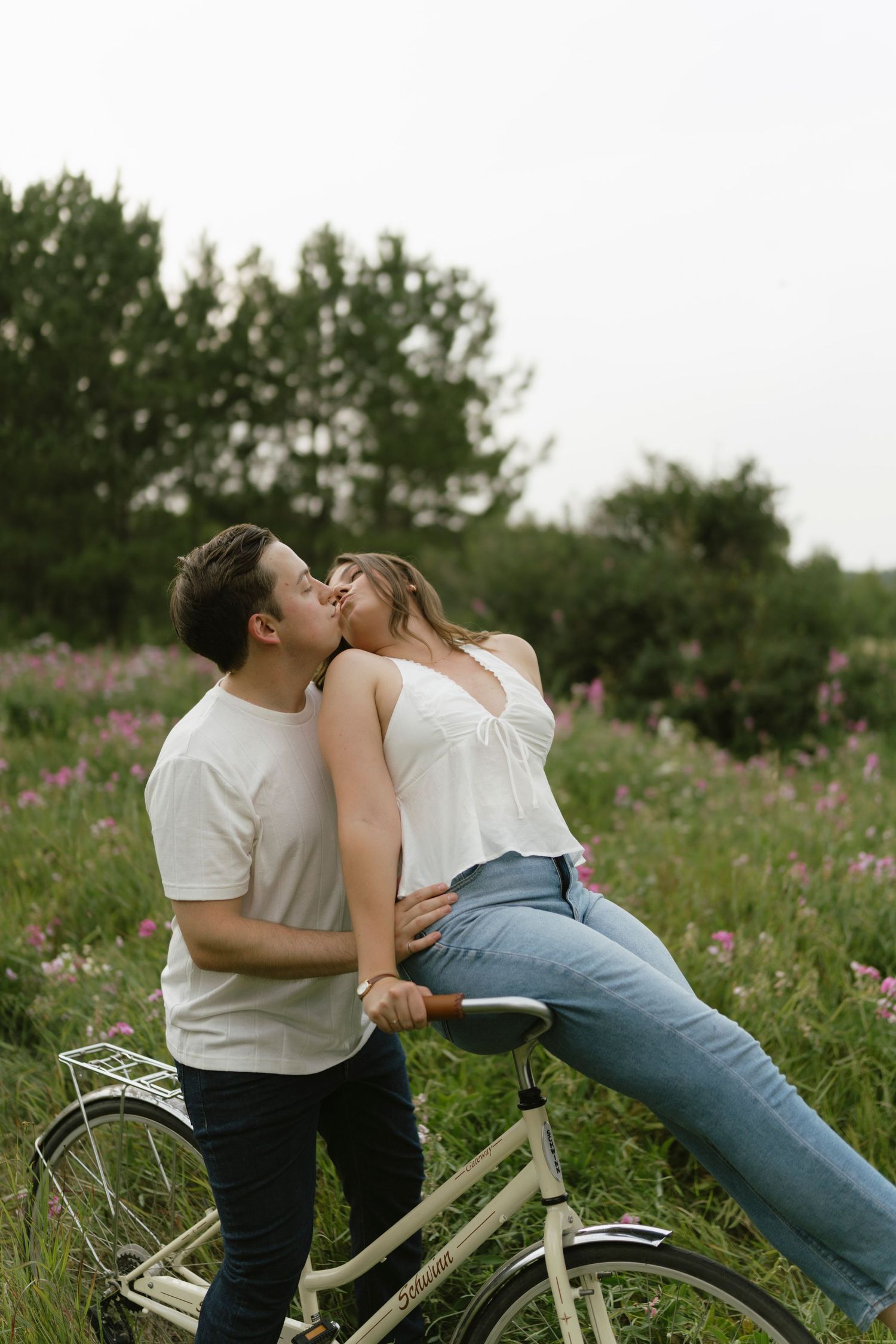 Couple sharing a romantic kiss outdoors, woman sitting on a vintage white bicycle surrounded by wildflowers and lush greenery.