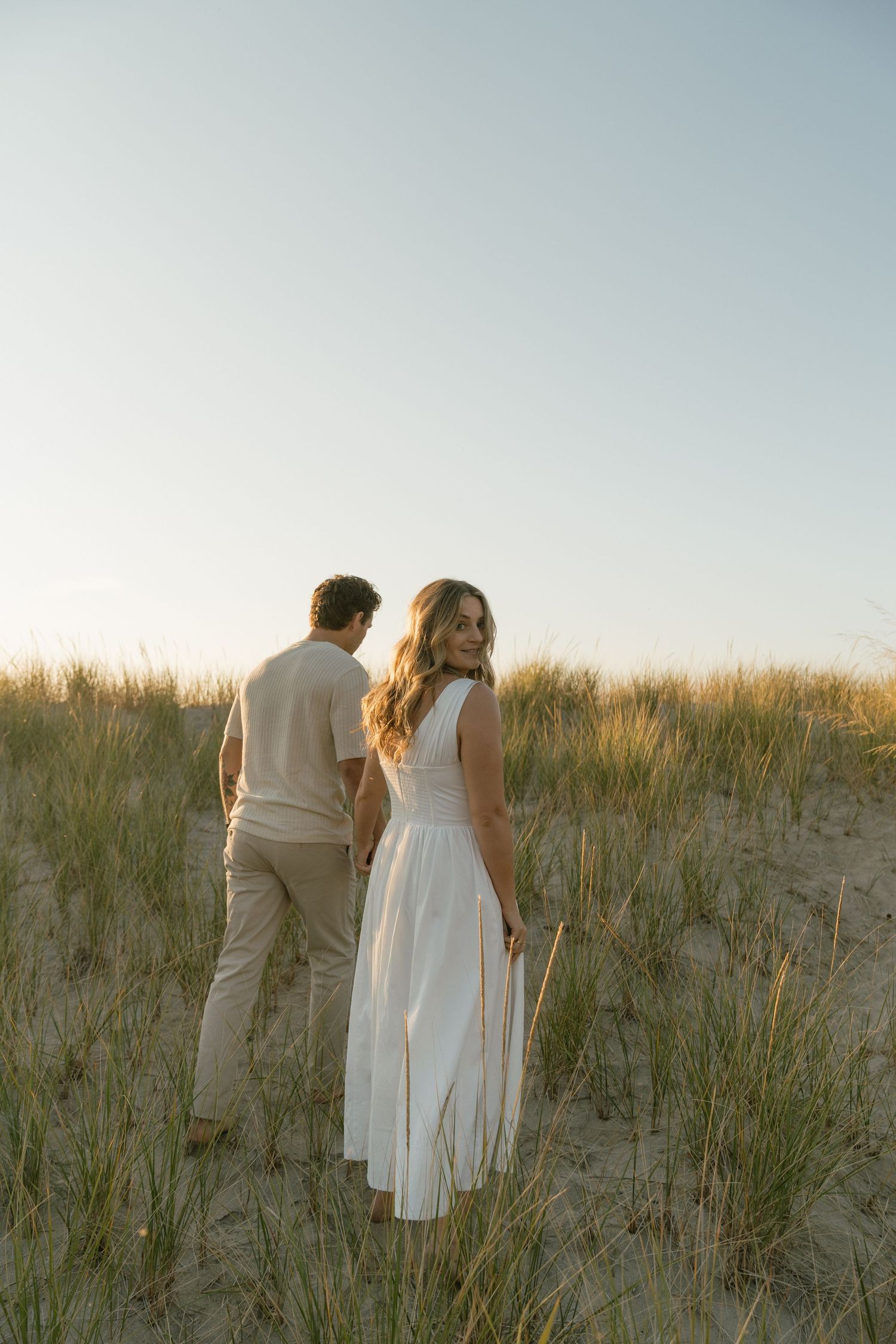 A couple in white outfits walking together through beach grass at sunset.