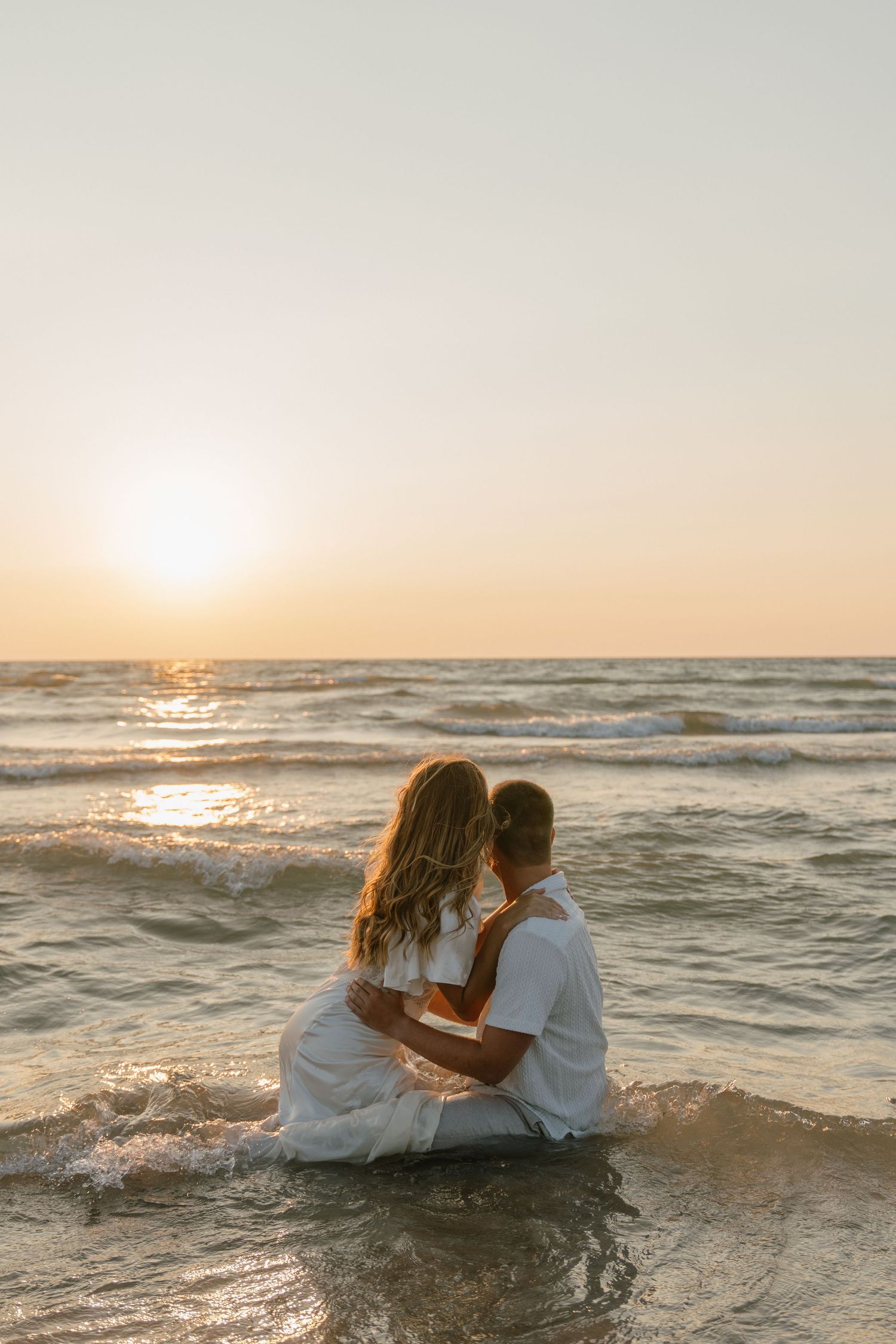 Couple sitting in ocean waves at sunset during romantic beach photoshoot.