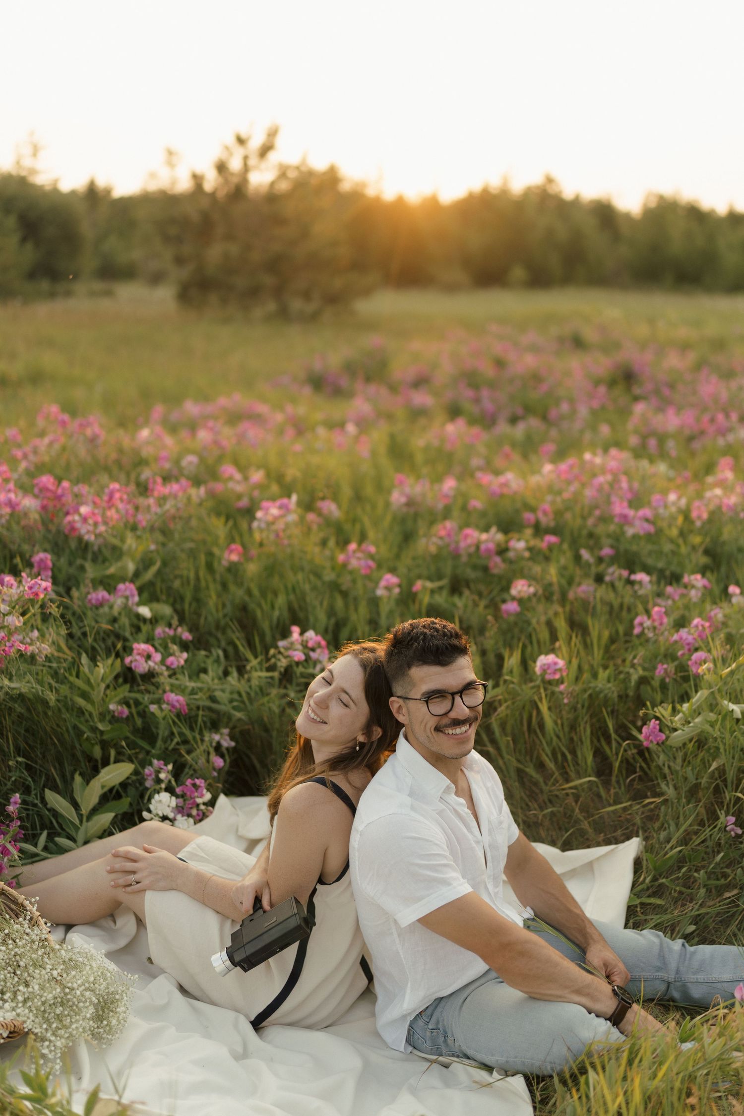 Couple relaxing on white blanket in field of pink wildflowers at golden hour.