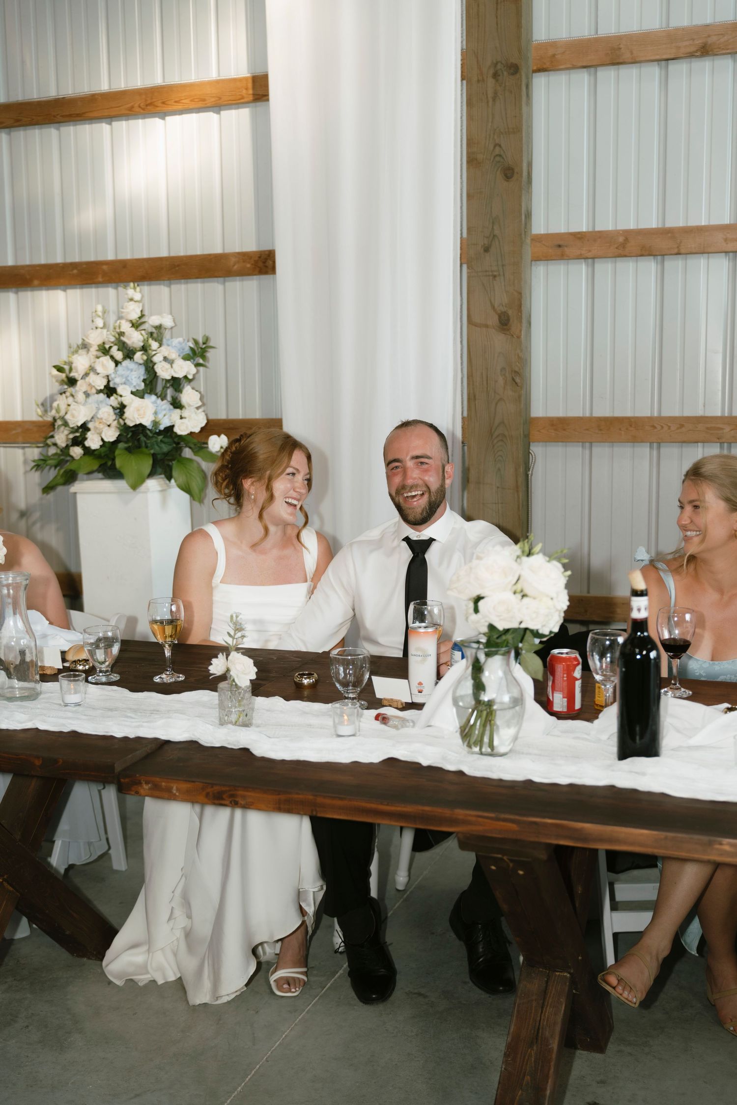 Bride and groom laughing at wedding reception head table in rustic barn venue with white floral arrangements.