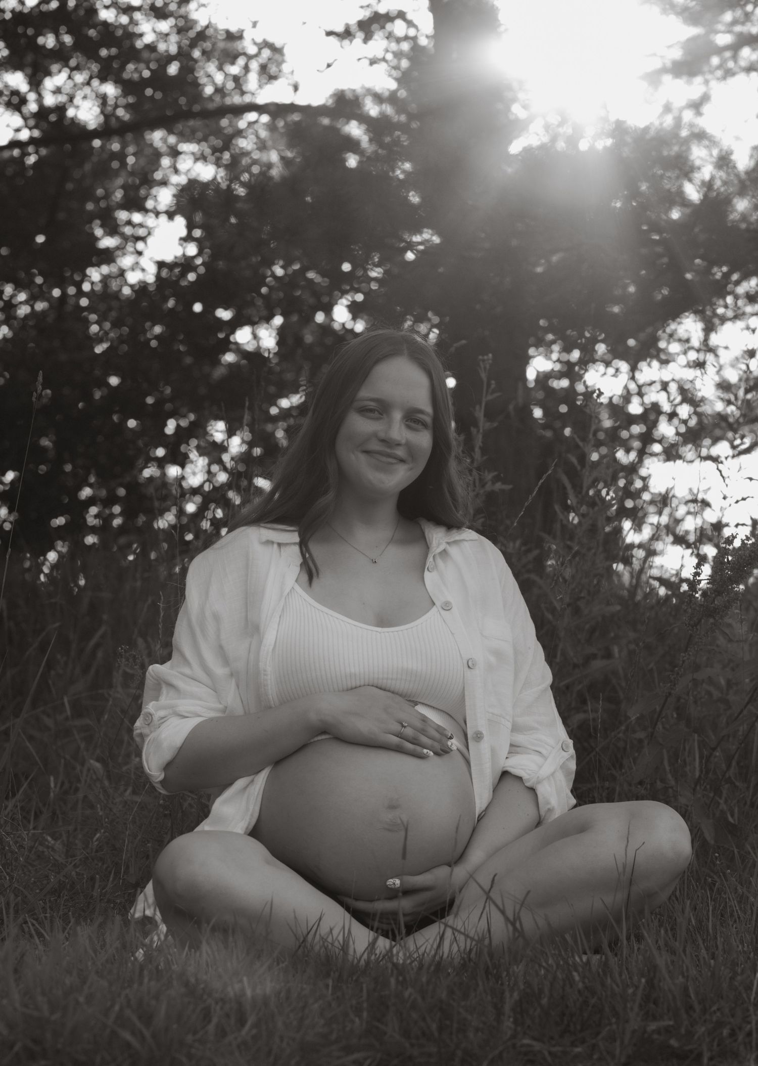 Black and white maternity photos showing a pregnant woman relaxing in grass during golden hour.