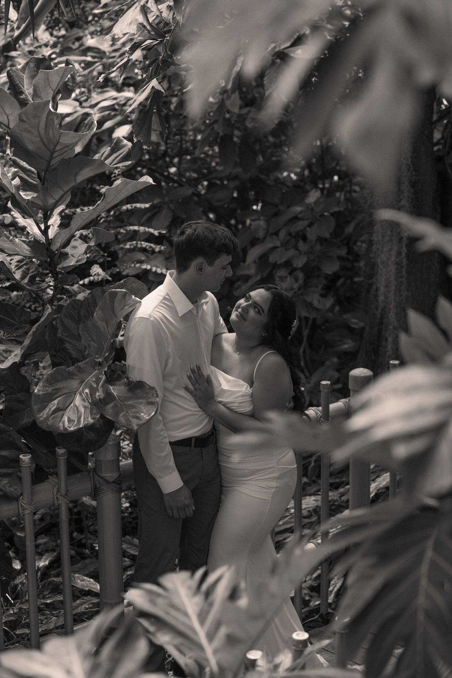 Black and white photo of a couple sharing tender moments against a leafy backdrop.