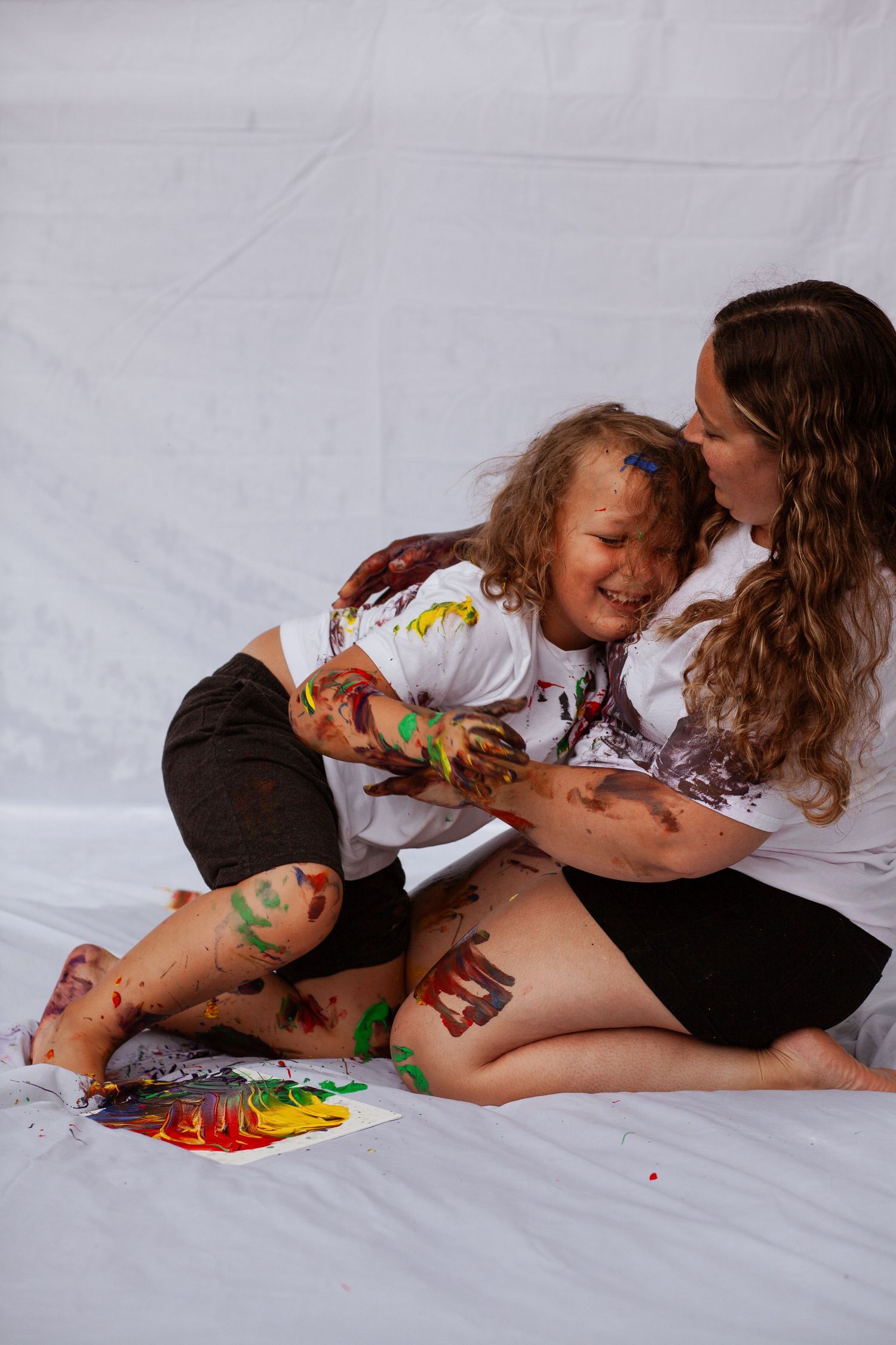 Two people playfully paint with colorful paints while lying on a white sheet.