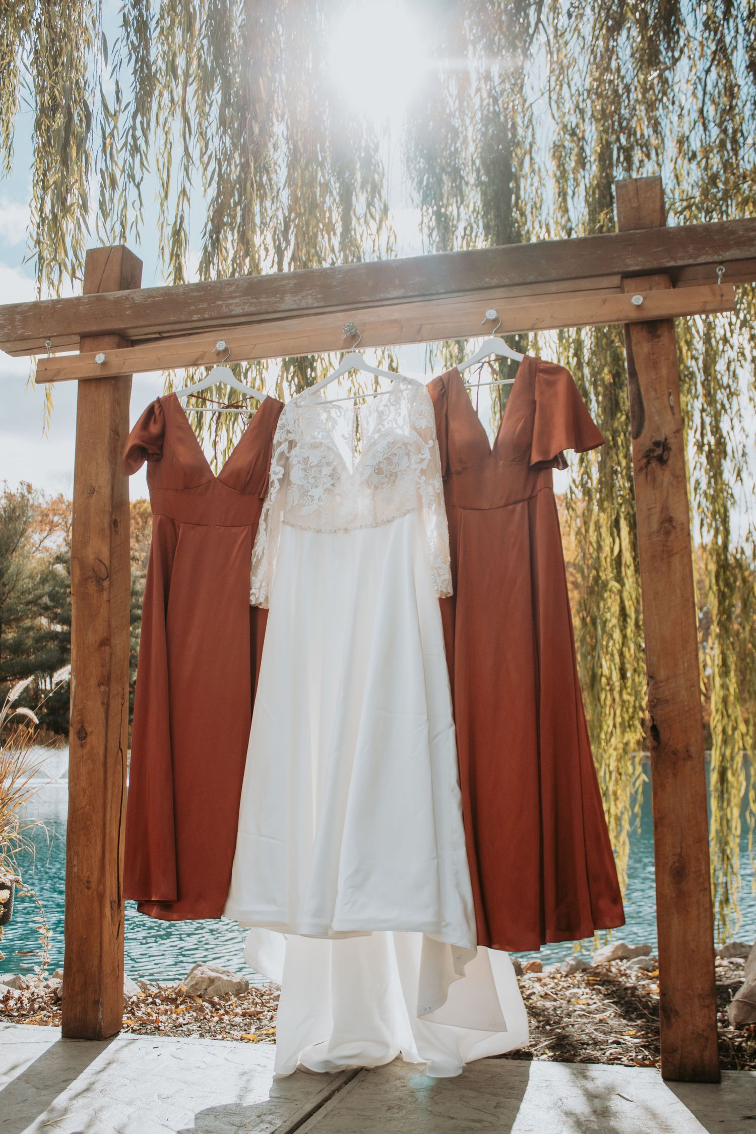 Wedding dresses and burgundy bridesmaid gowns hanging on wooden pergola by water.