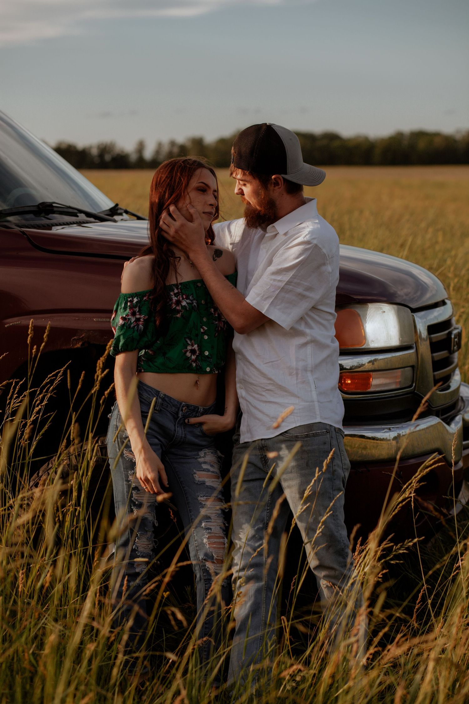 Romantic couple posing by red pickup truck in a rustic field at sunset.
