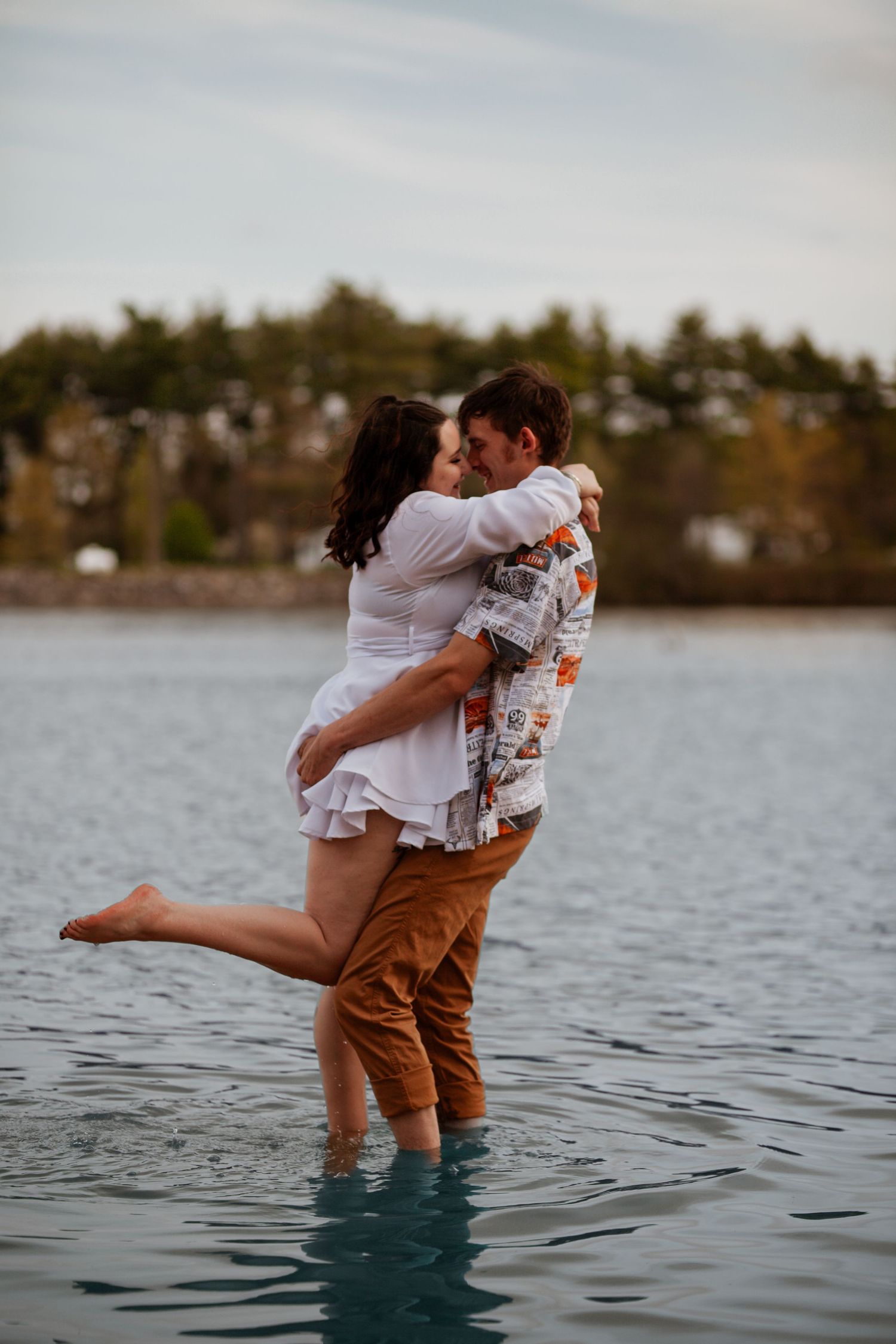 Playful couple embracing while standing in water with scenic landscape behind them.