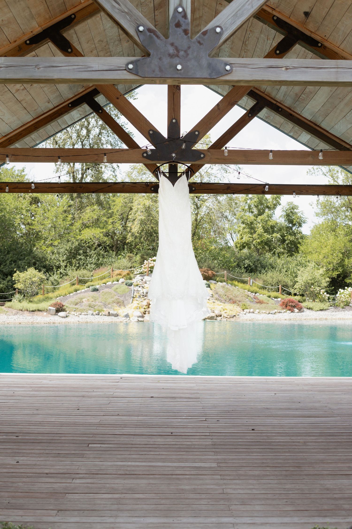A white wedding dress hangs from a wooden pavilion overlooking a serene turquoise pool and landscaped garden.