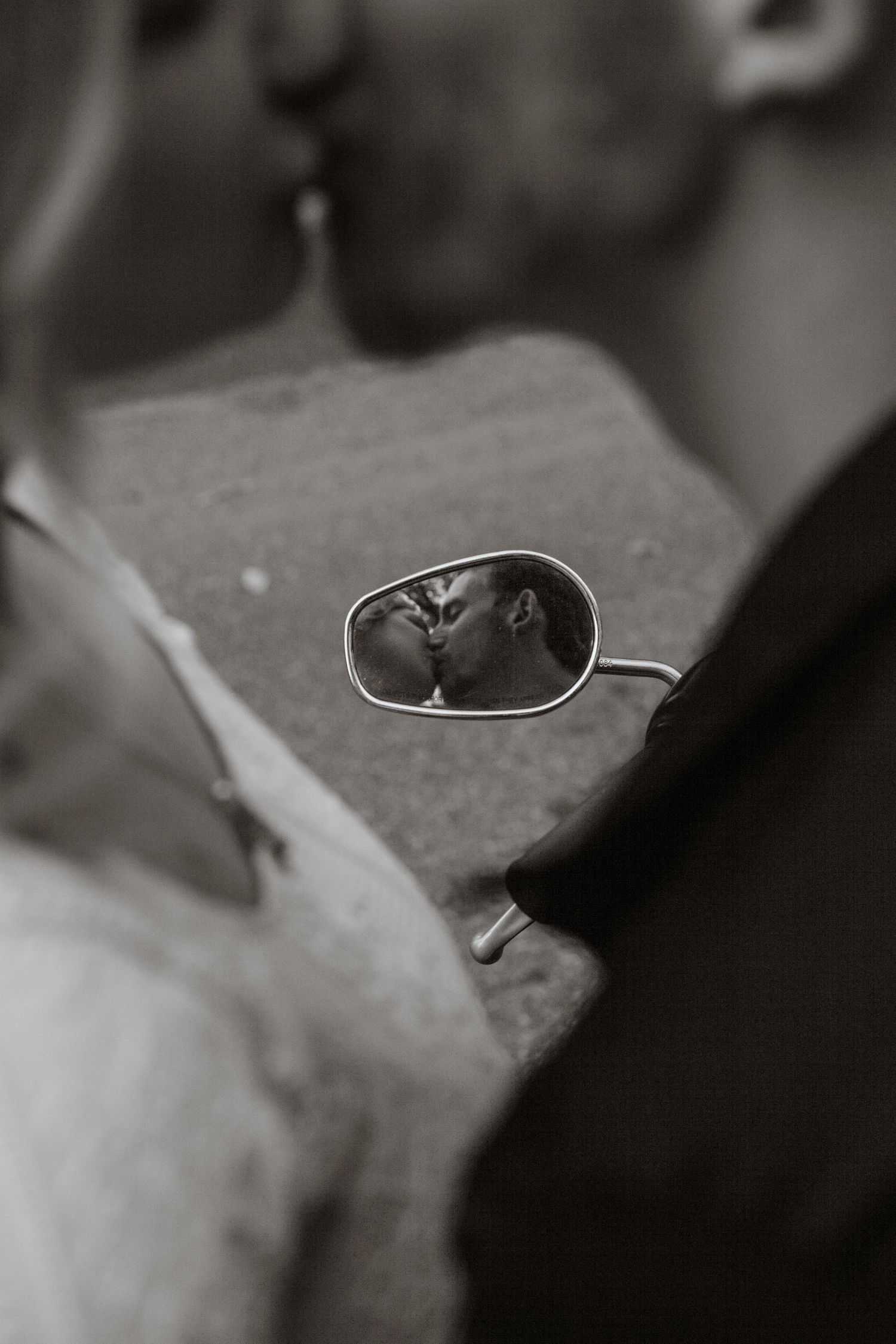 A creative black and white photo showing a couple's reflection in a motorcycle mirror.