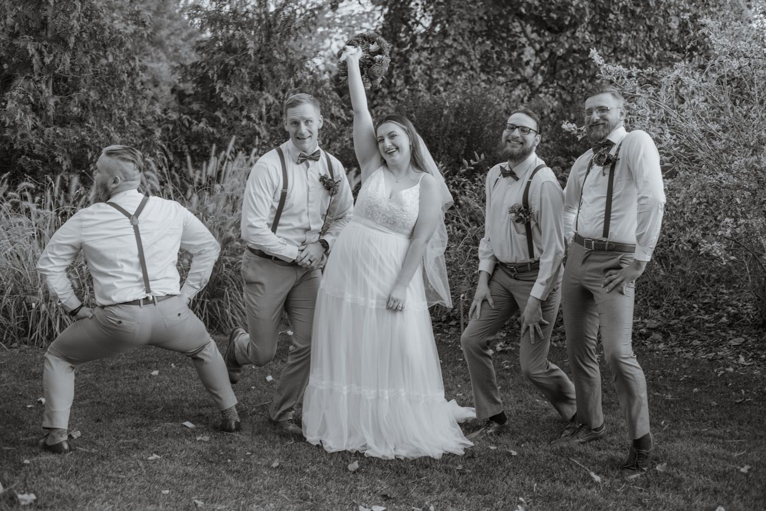 Wedding party poses playfully in a garden setting with suspenders and formal attire.