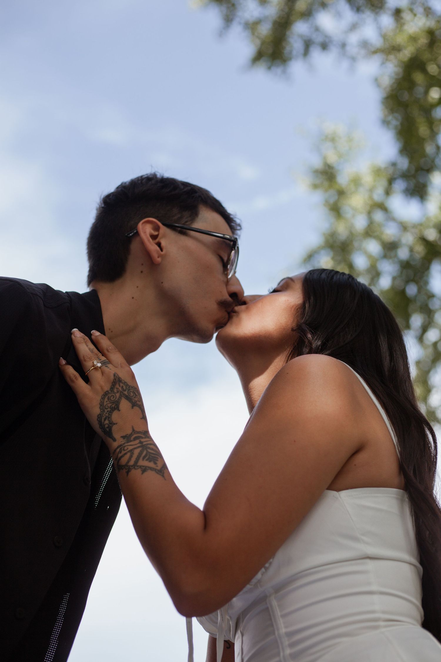 Romantic outdoor photoshoot with couple kissing against blue sky and trees.