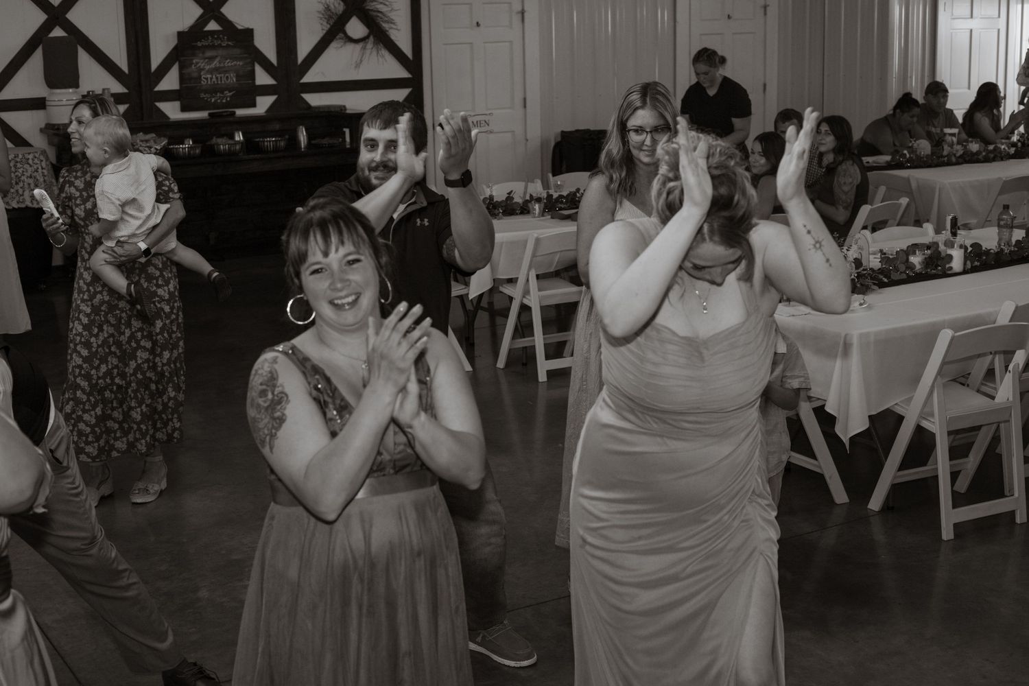 Black and white photo of wedding guests dancing and celebrating at an indoor reception.