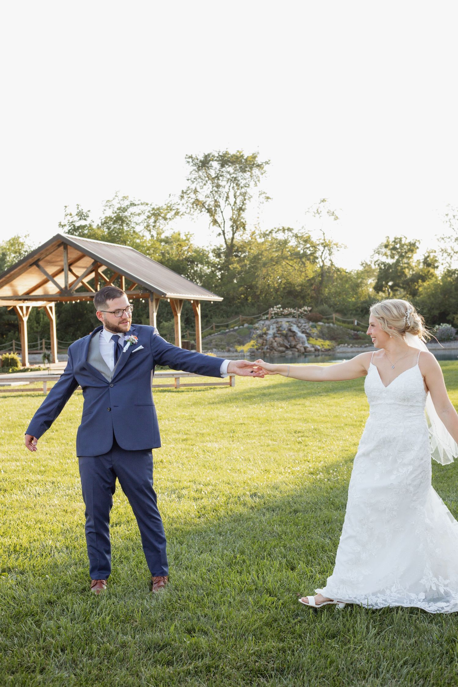 A couple in wedding attire holding hands while standing apart on a sunny lawn with a gazebo in the background.