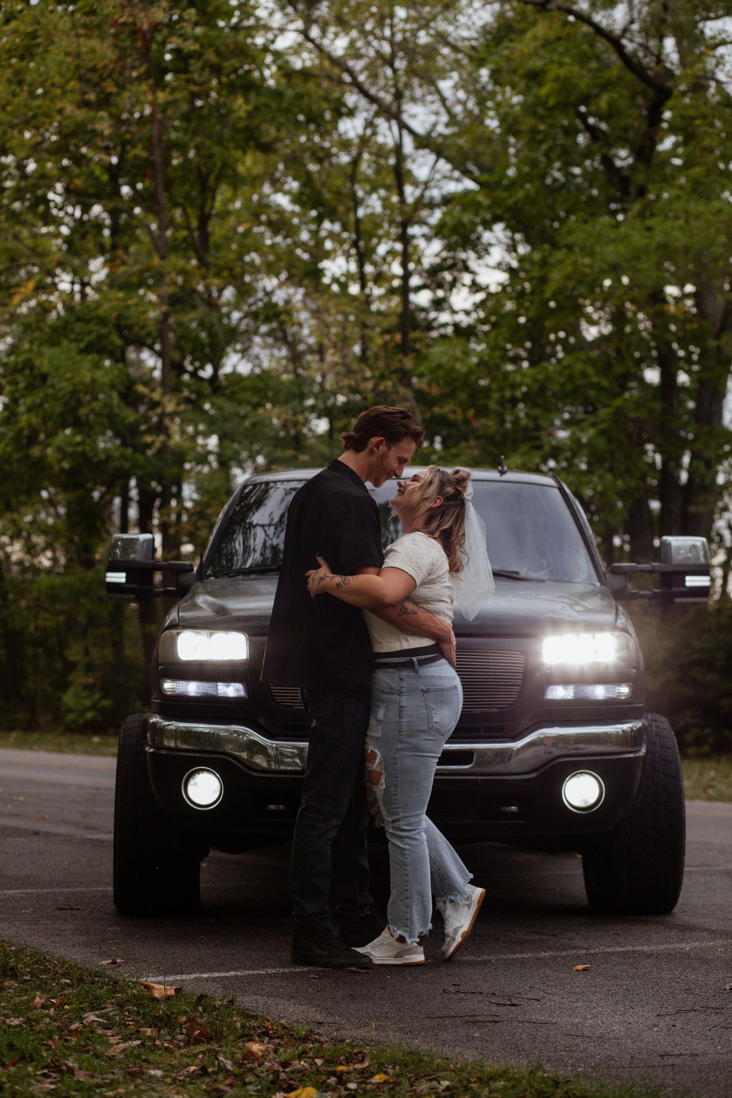 Couple embracing in front of cars with headlights on at dusk.