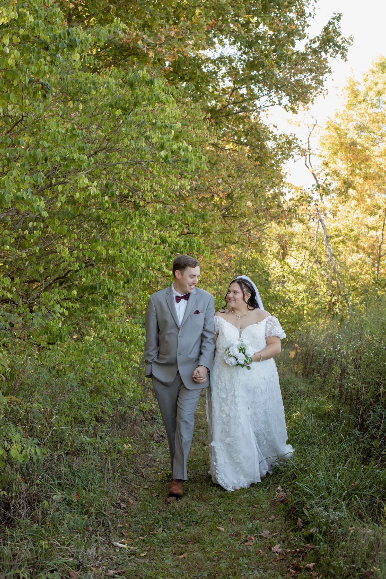 Couple in wedding attire walking through a sunlit wooded path.