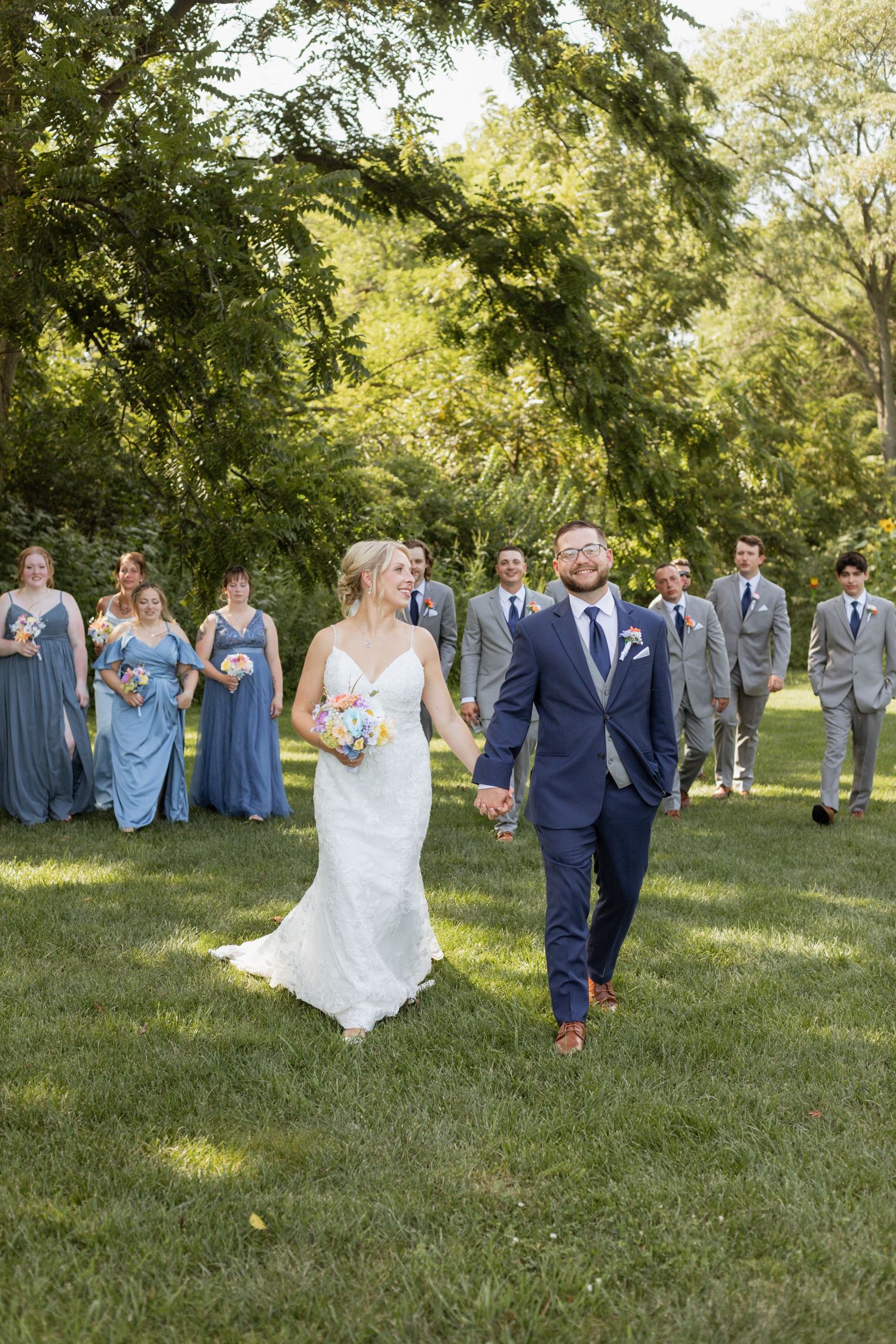 Wedding couple walking through a sun-dappled garden with bridal party in blue dresses.