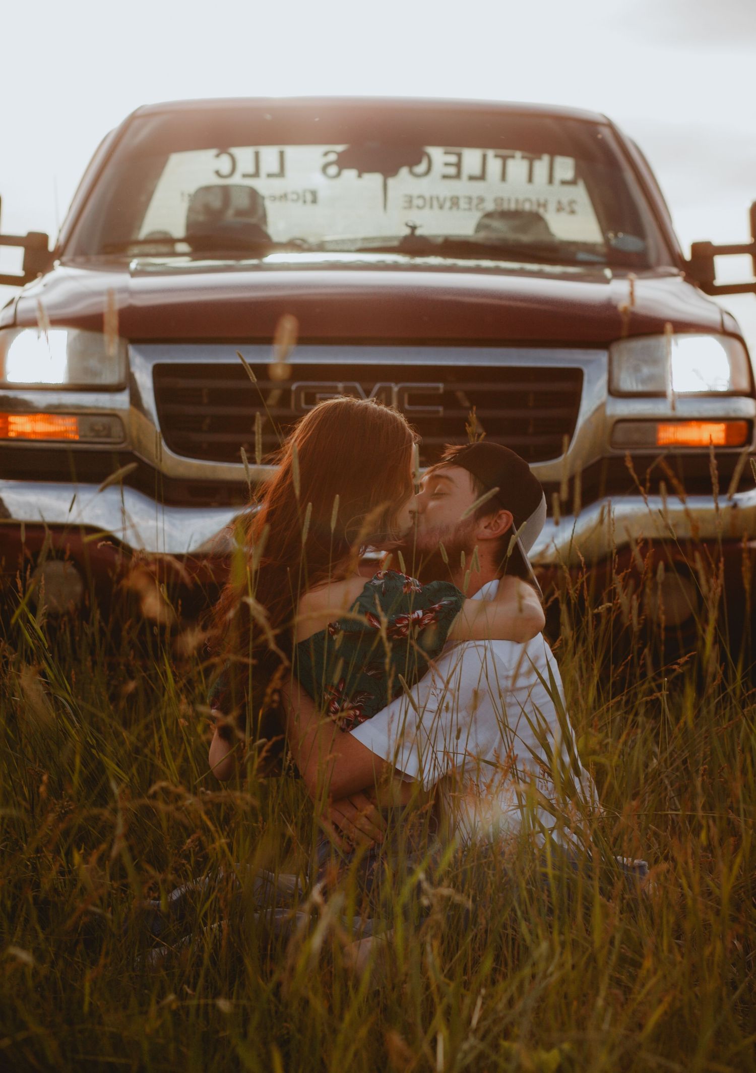 Couple embracing in tall grass with a pickup truck in the background during golden hour.