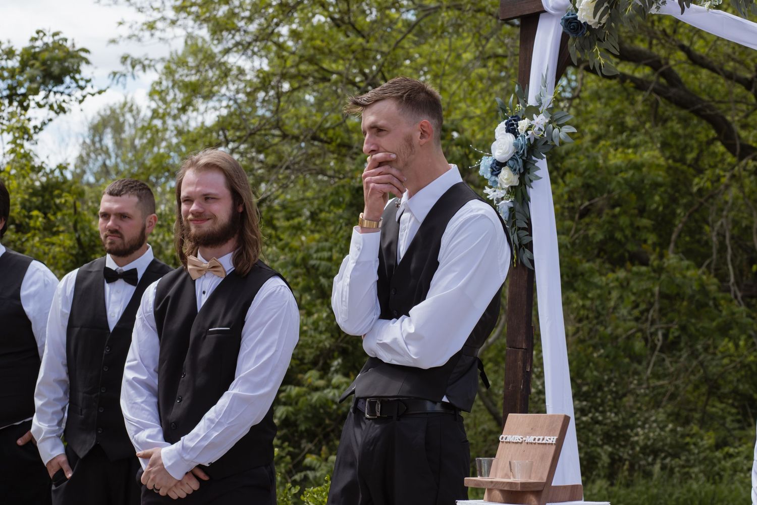 Groomsmen in vests and ties standing at an outdoor wedding ceremony.