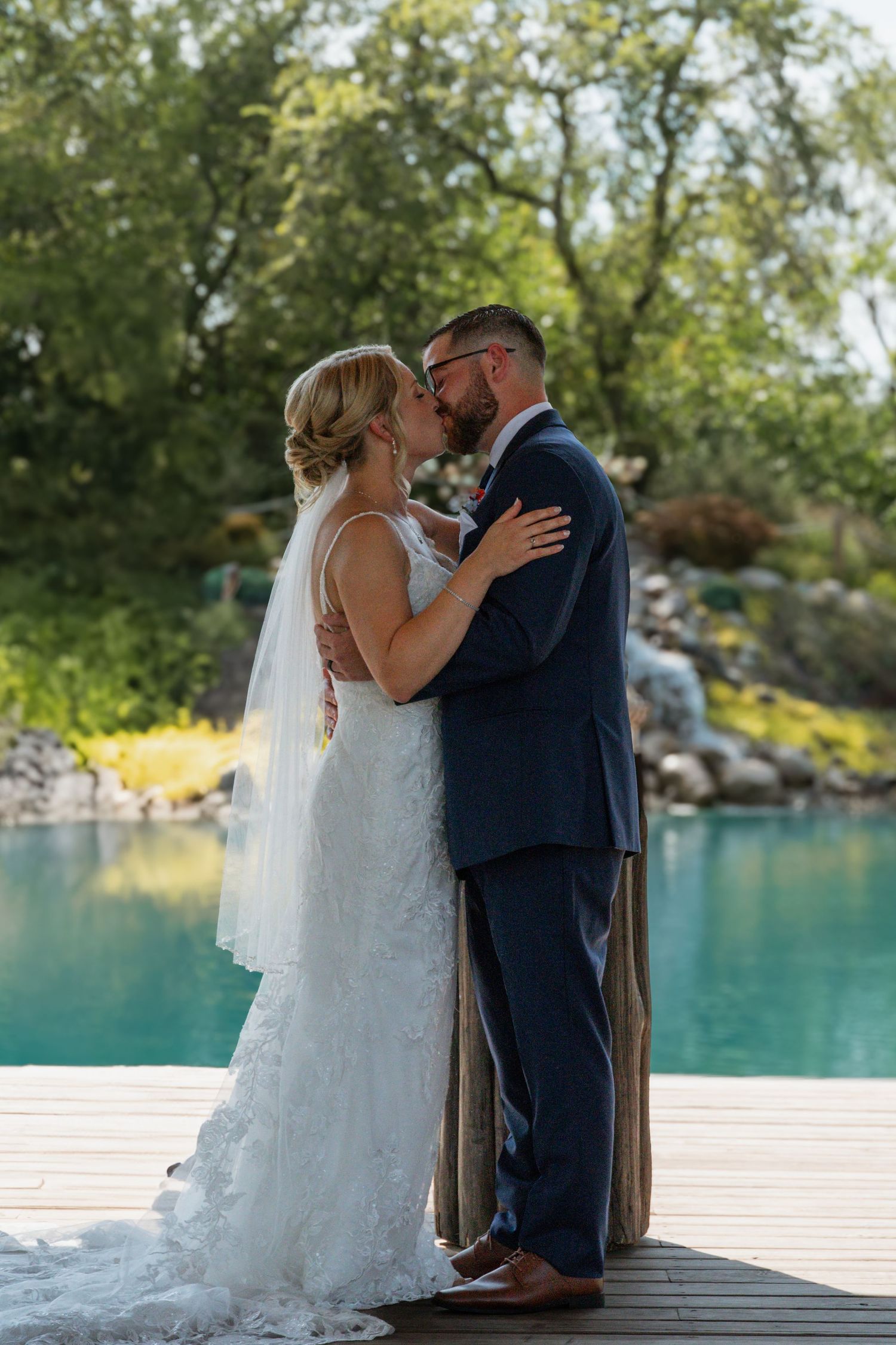 Romantic wedding kiss on wooden deck overlooking turquoise pool and garden landscape.