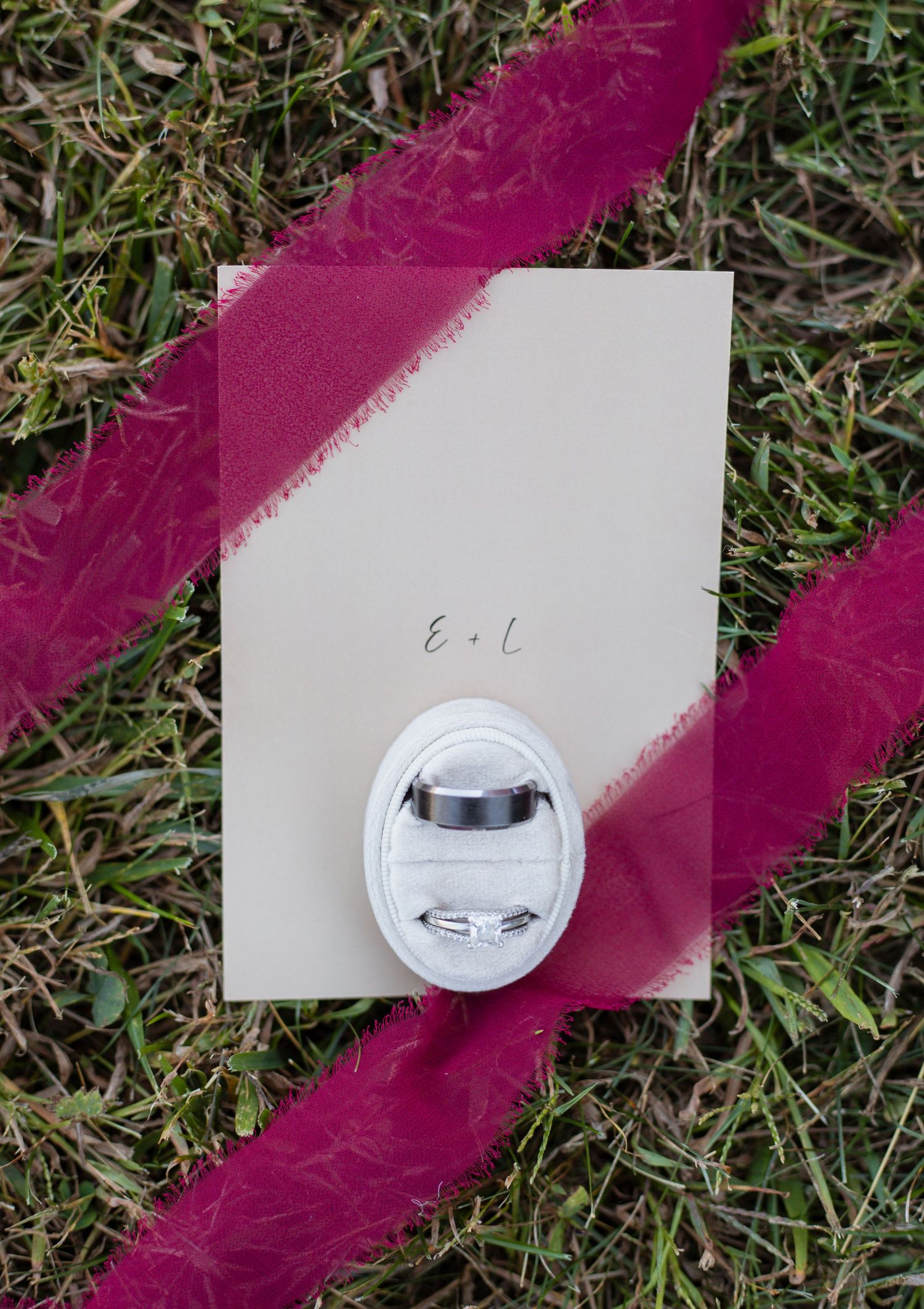 White wedding card with decorative pin placed on grass with magenta ribbon accent.