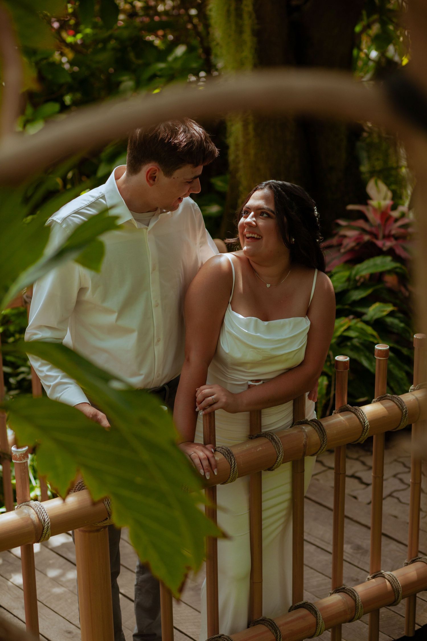 Romantic couple in white attire share intimate moments along a wooden railing in a garden setting.