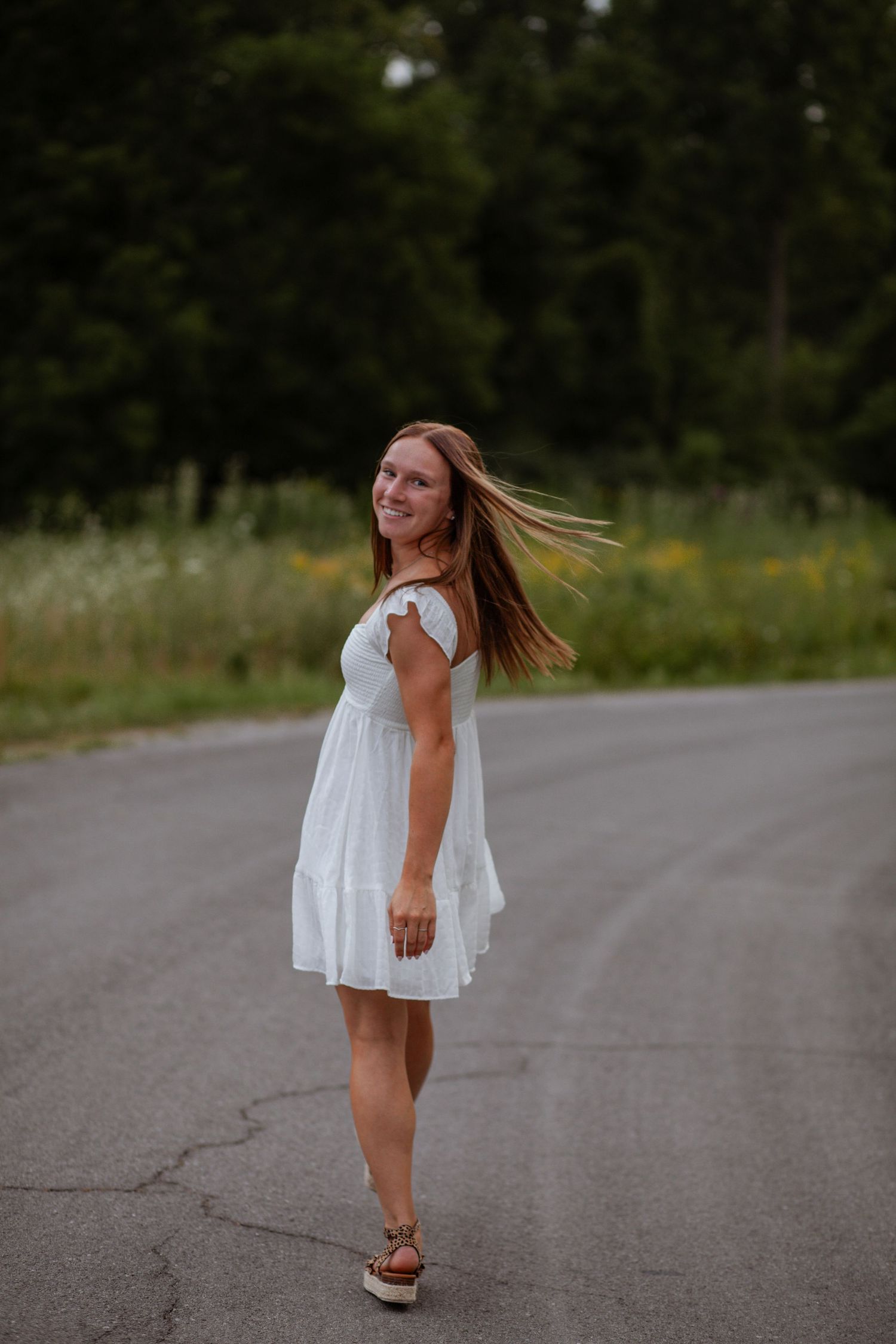 A figure in a flowing white dress twirls and dances on a rural road at sunset.