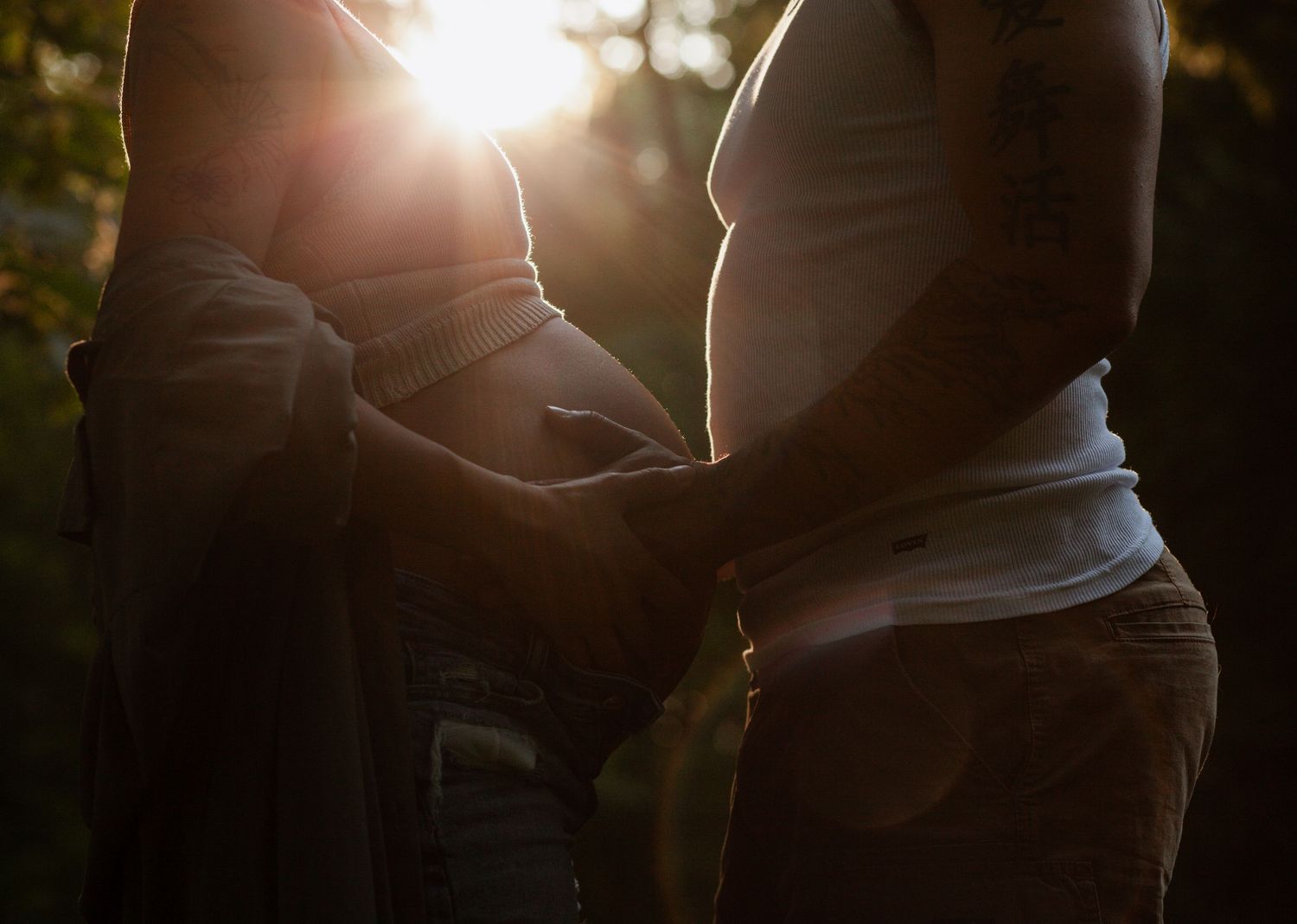 Silhouetted maternity photo of two people touching a pregnant belly in golden sunset light.