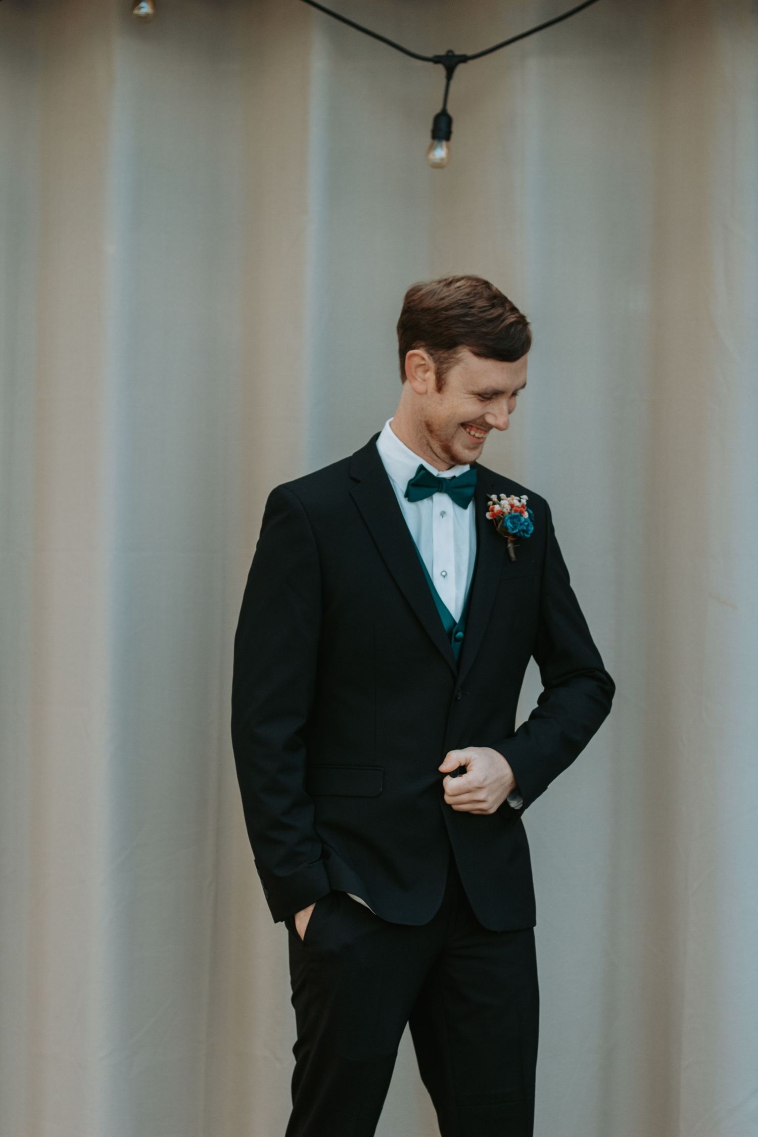 Groom in classic black tuxedo and bowtie standing against light curtain backdrop.
