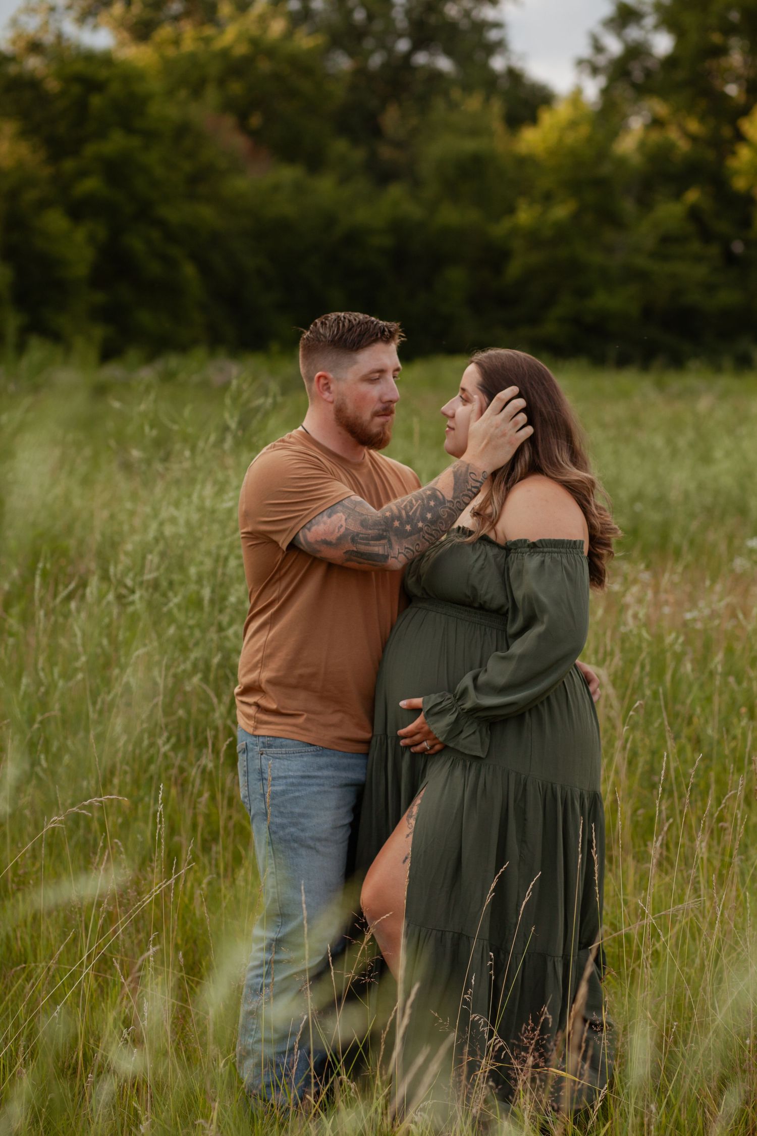 A romantic couple embraces in a sunlit meadow with tall grass and trees in the background.