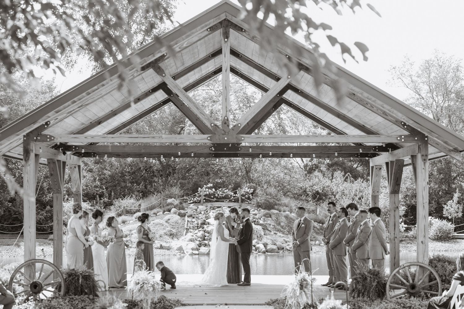 Black and white wide shot of wedding ceremony under rustic wooden pavilion with gathered guests.