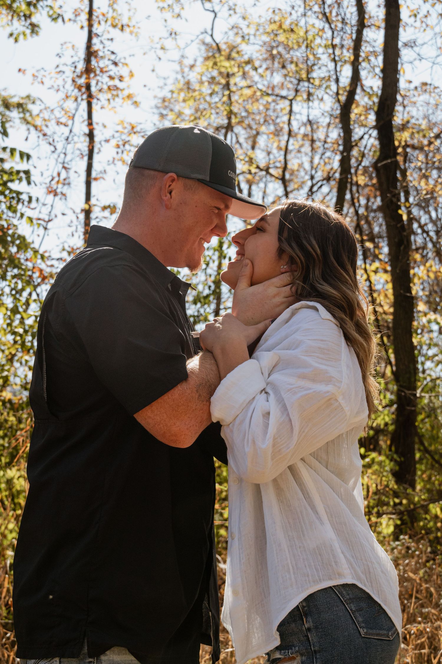A couple embracing in sunlit woods wearing casual attire during an autumn day.